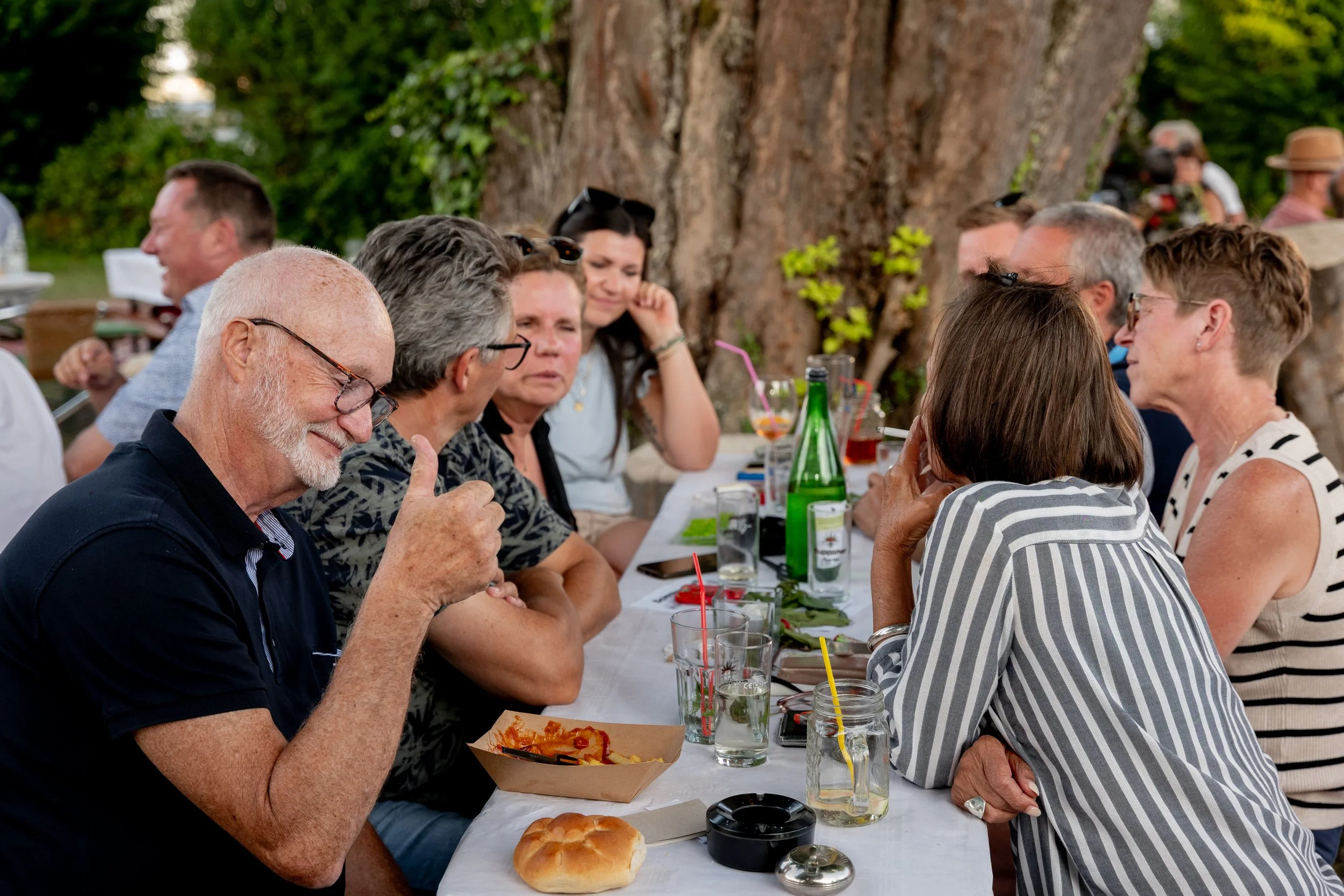 Menschen sitzen an einem langen Tisch im Freien, essen und trinken, bei einer Gartenfeier oder einem Treffen, mit Bäumen im Hintergrund.