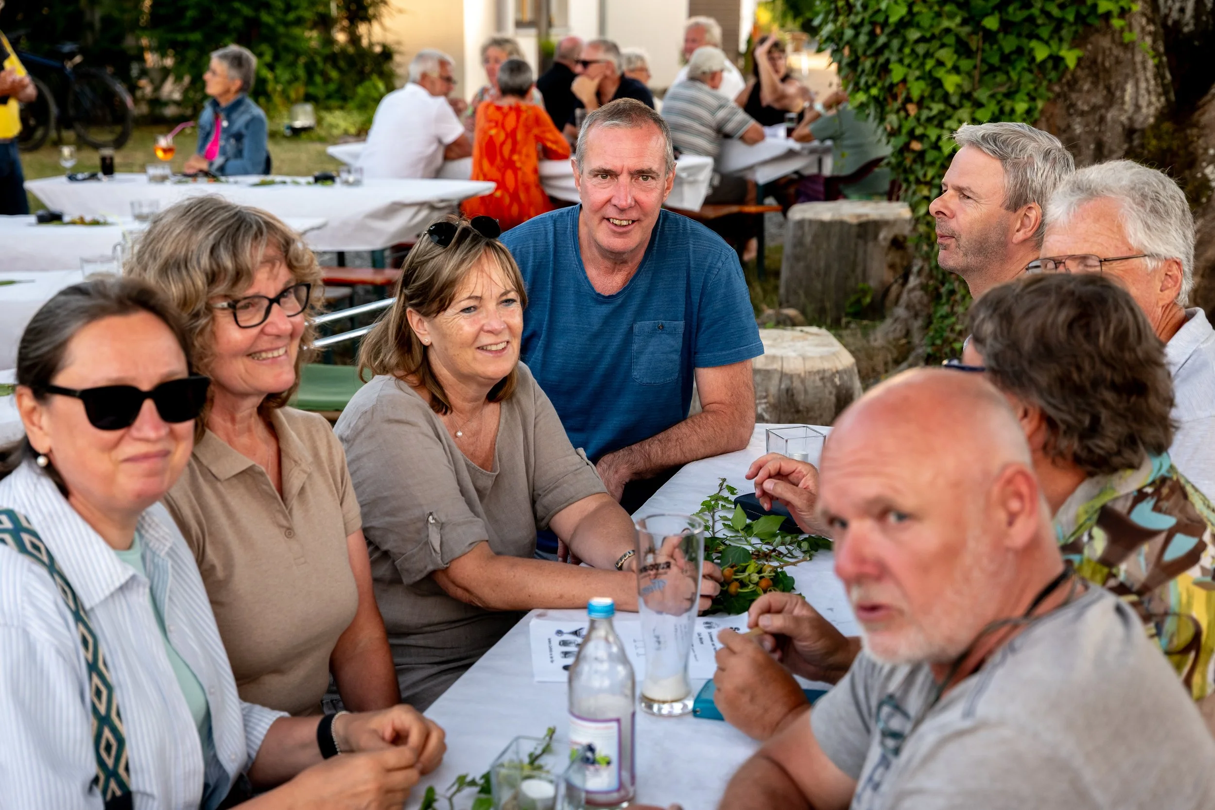 Menschen sitzen zusammen an einem langen Tisch im Freien bei einer Gartenparty, trinken Getränke und unterhalten sich.