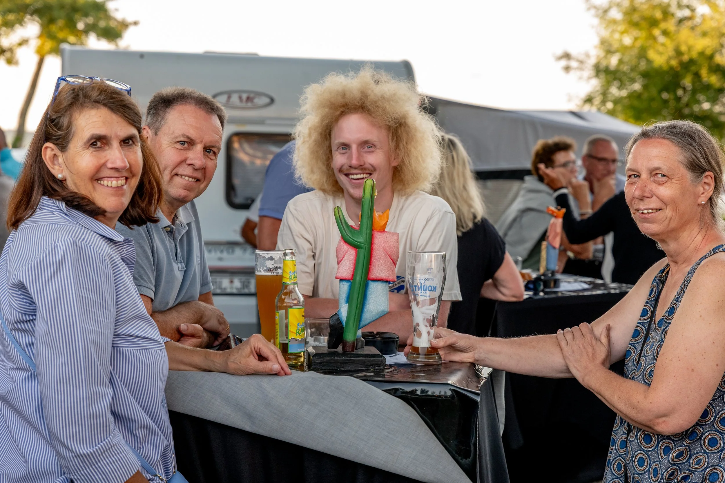 Menschen sitzen an einem Tisch im Freien bei einem Treffen, trinken Bier, lachen und haben Spaß, mit einem Wohnwagen im Hintergrund.
