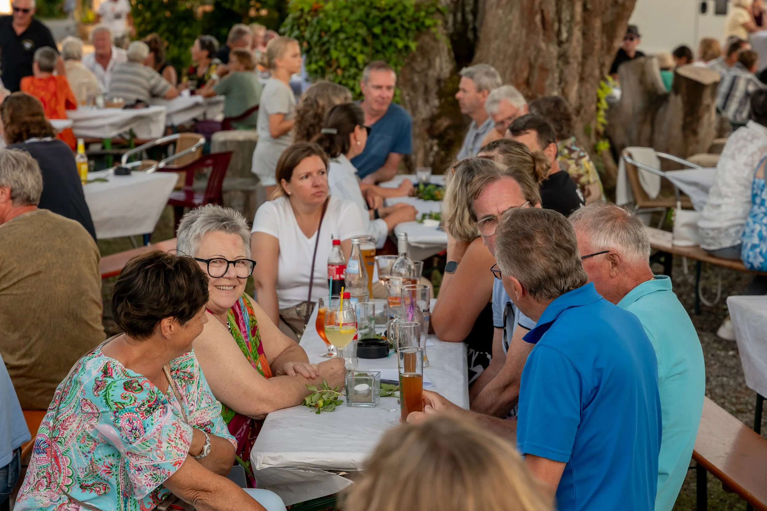 Menschen sitzen bei einer Outdoor-Party an Tischen, trinken Getränke und unterhalten sich, mit Bäumen und mehr Menschen im Hintergrund.