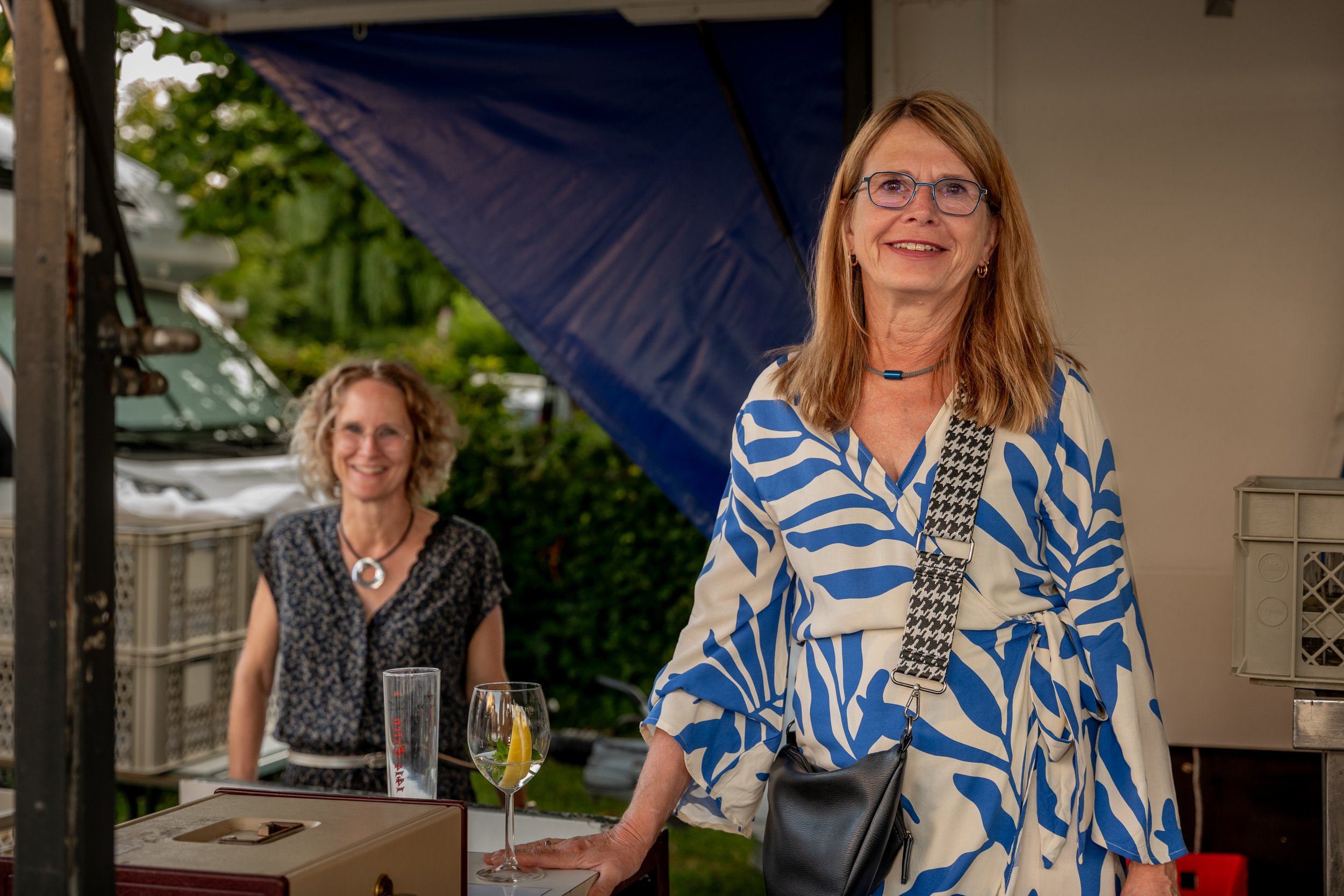 Zwei Frauen bei einer Gartenparty, eine steht am Tisch und lächelt in die Kamera, die andere sitzt im Hintergrund unter einem blauen Sonnenschirm mit einem Getränk und einem Halskett, im Freien mit Bäumen und einem Anhänger im Hintergrund.
