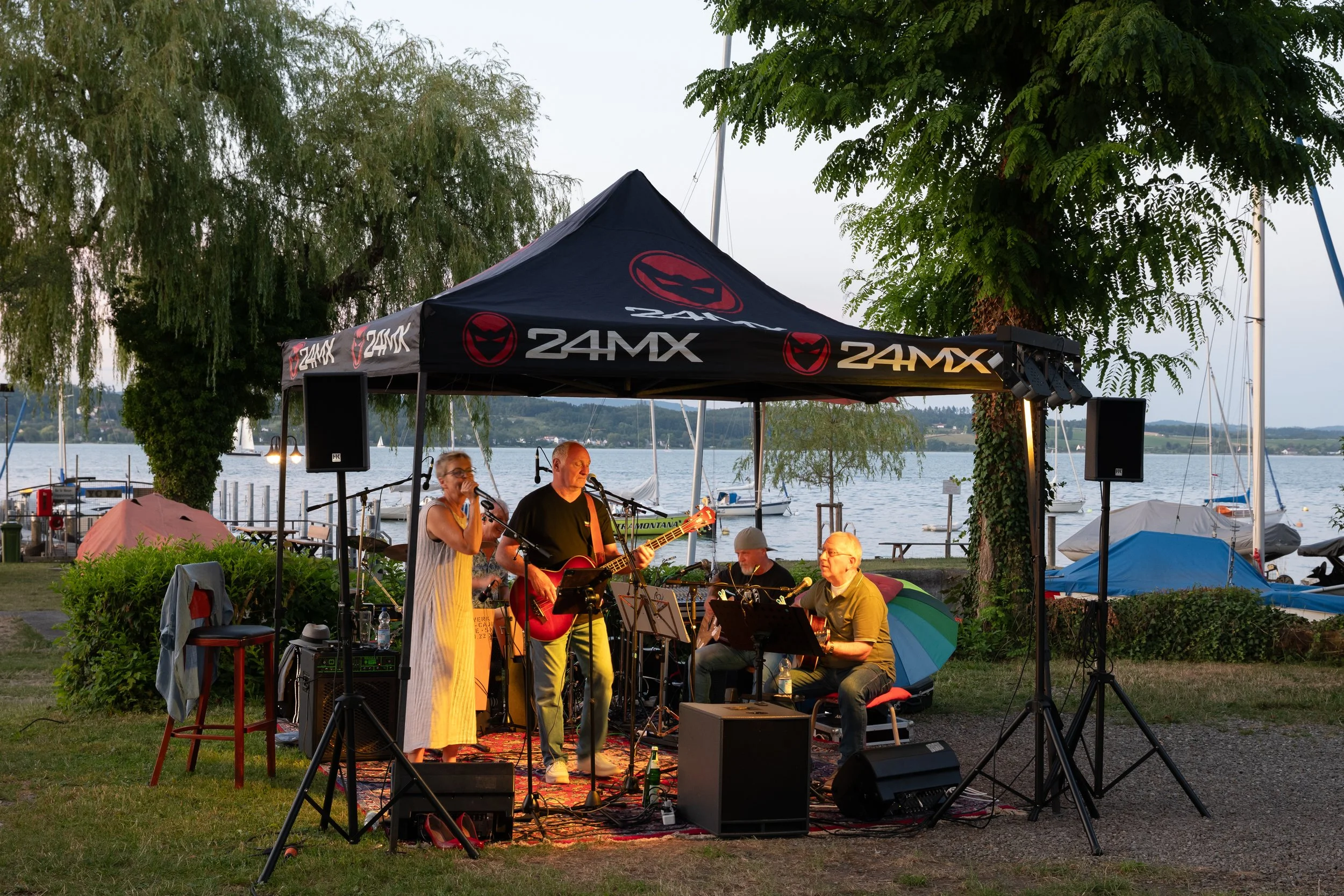 Musiker spielen bei einem Open-Air-Konzert am See unter einem schwarzen Zelt mit rotem Logo und Schriftzug "24MX". Es sind Menschen mit Gitarren und Instrumenten, im Hintergrund sind Boote im Wasser zu sehen.