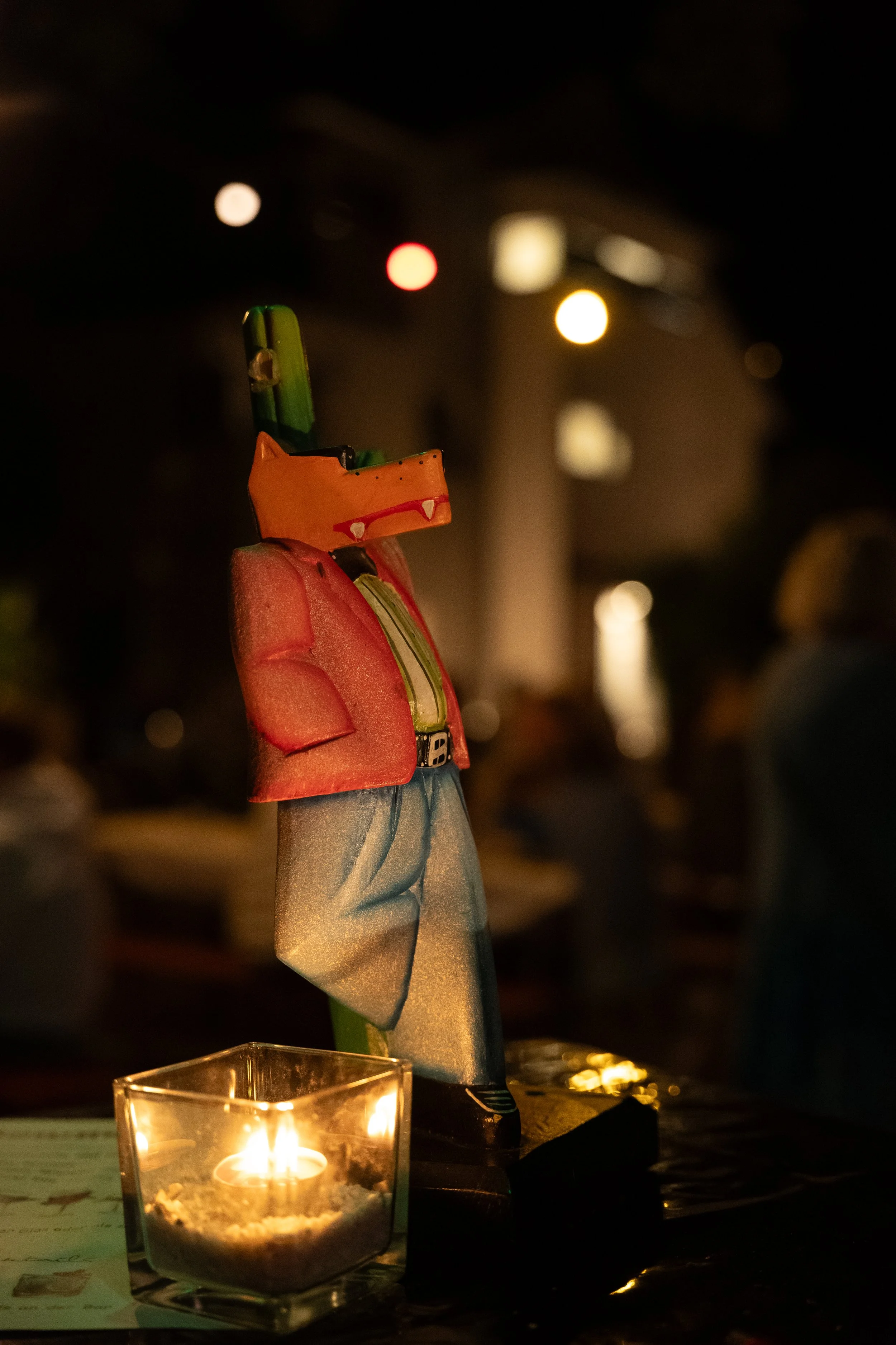 Papierfigur eines Ansprechpartners in einem pinken Jacke, grauen Hosen und schwarzen Schuhen, auf einem Tisch mit einer Kerze in einem Glas. Hintergrund unscharf, dimmige Beleuchtung.