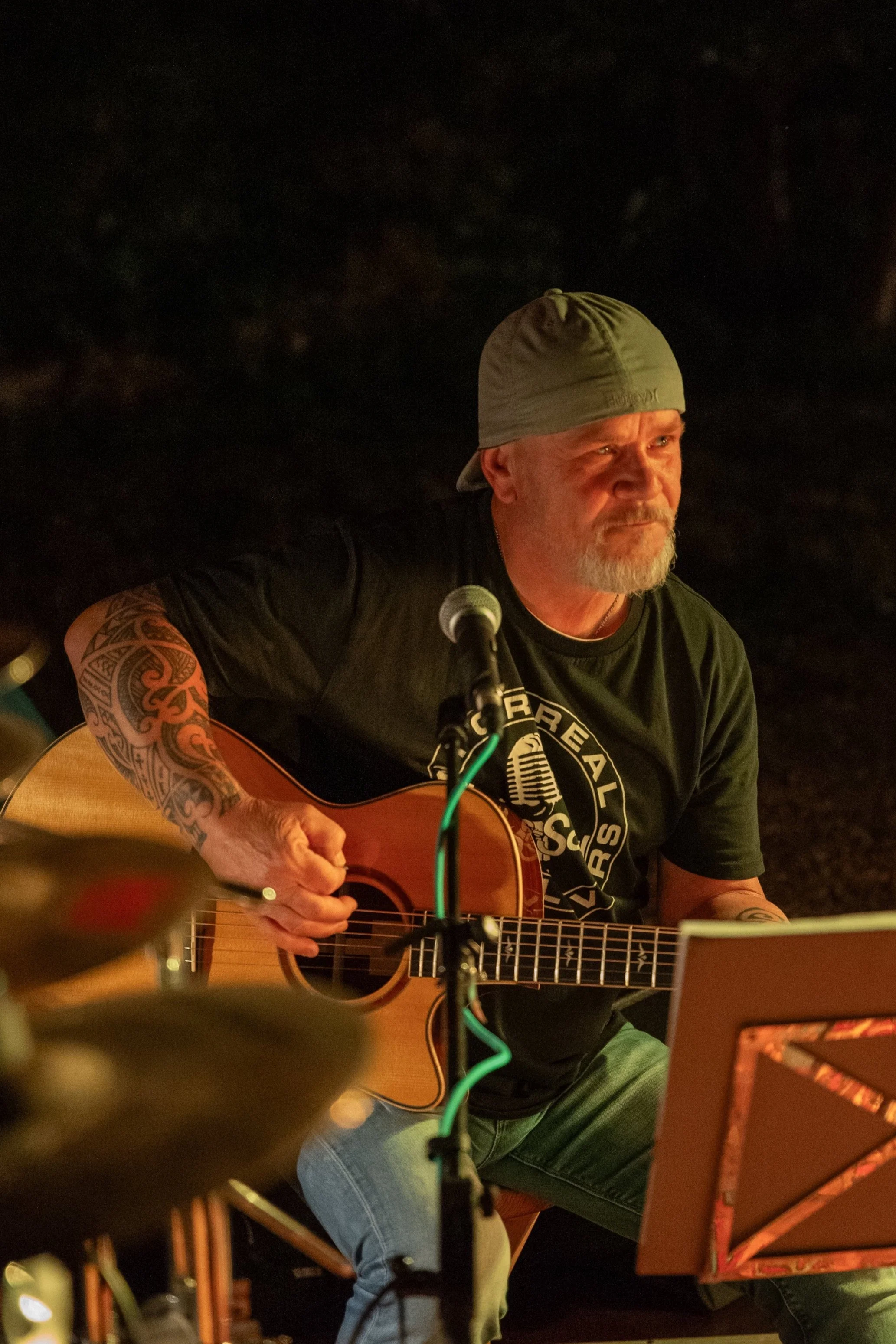 Ein Mann spielt Gitarre bei einer Live-Musikveranstaltung, trägt ein graues Cap und ein schwarzes T-Shirt, sitzt bei Kerzenlicht in einer dunklen Umgebung.
