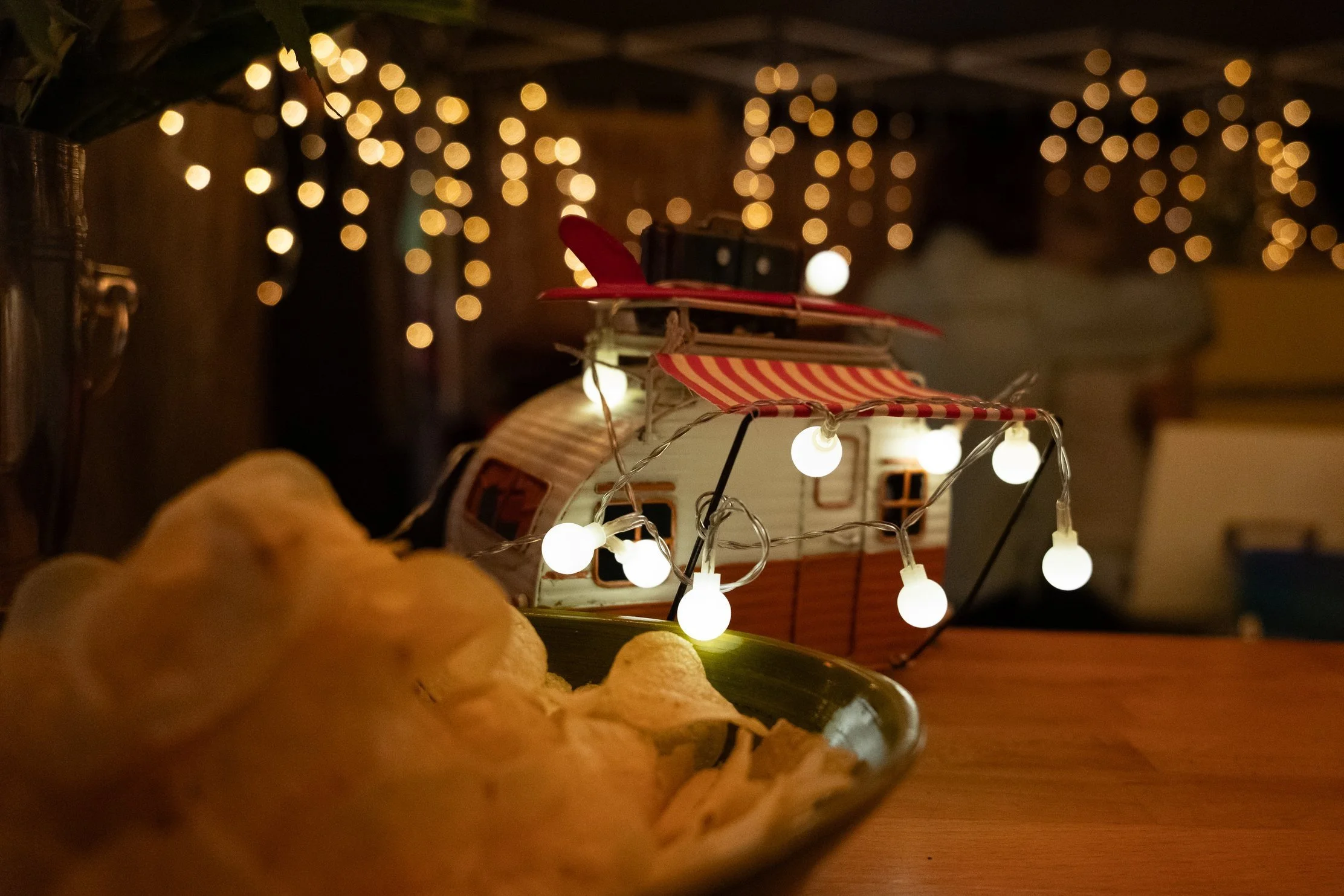 Eine im Dunkeln beleuchtete Weihnachtsdekoration mit einem Miniatur-Wohnmobil und einer Lichterkette. Im Vordergrund liegt eine Schale mit Chips. Im Hintergrund sind bunte Lichter und Dekorationen sichtbar.