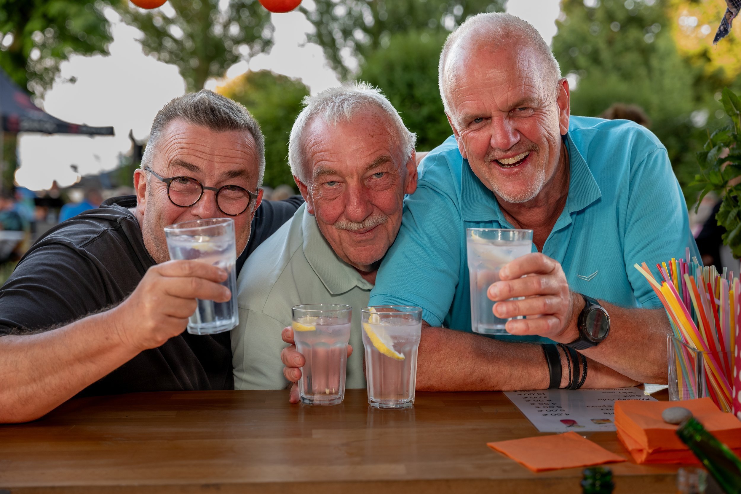 Drei Männer sitzen an einem Tisch im Freien, lachen und halten Gläser mit Wasser, Zitronenscheiben und Eis. Es ist eine fröhliche Atmosphäre bei einem Picknick oder Straßenfest.