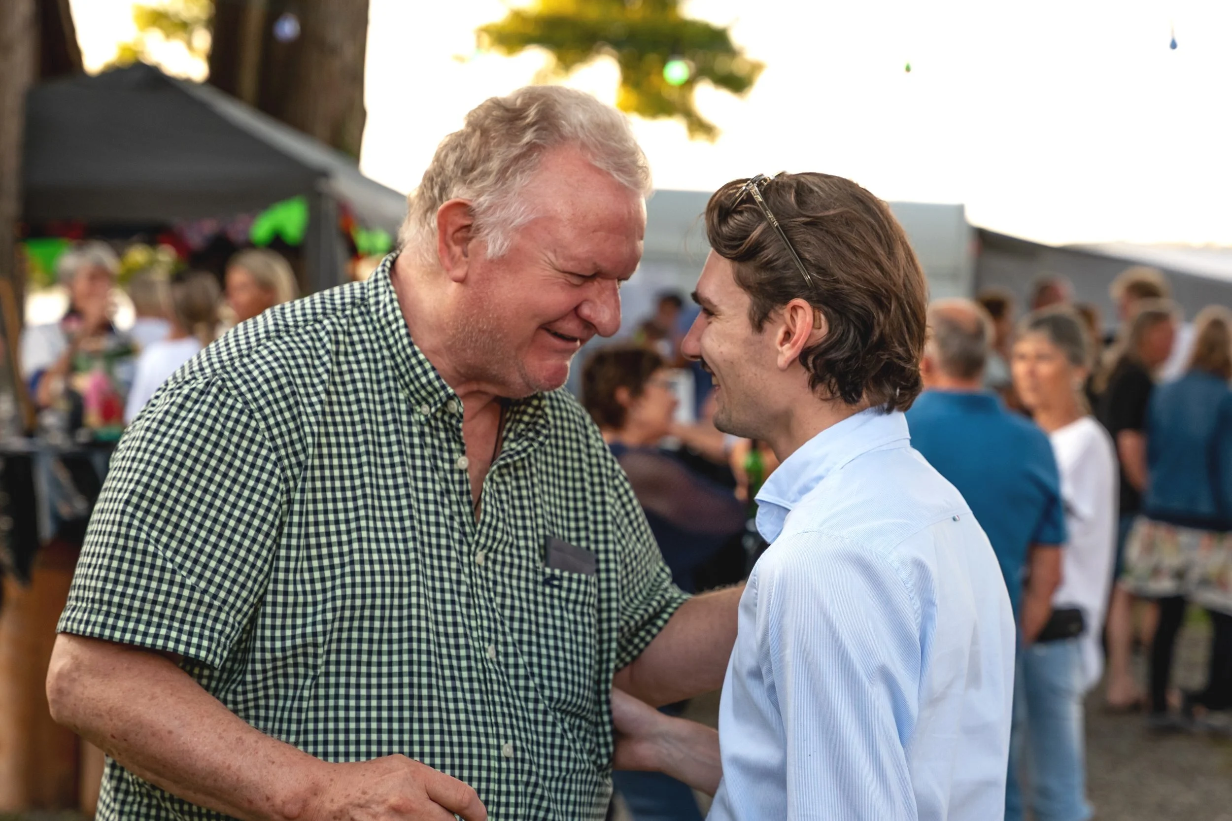 Zwei Männer, die auf einer Feier lachen und sich berühren, umgeben von anderen Menschen im Freien.