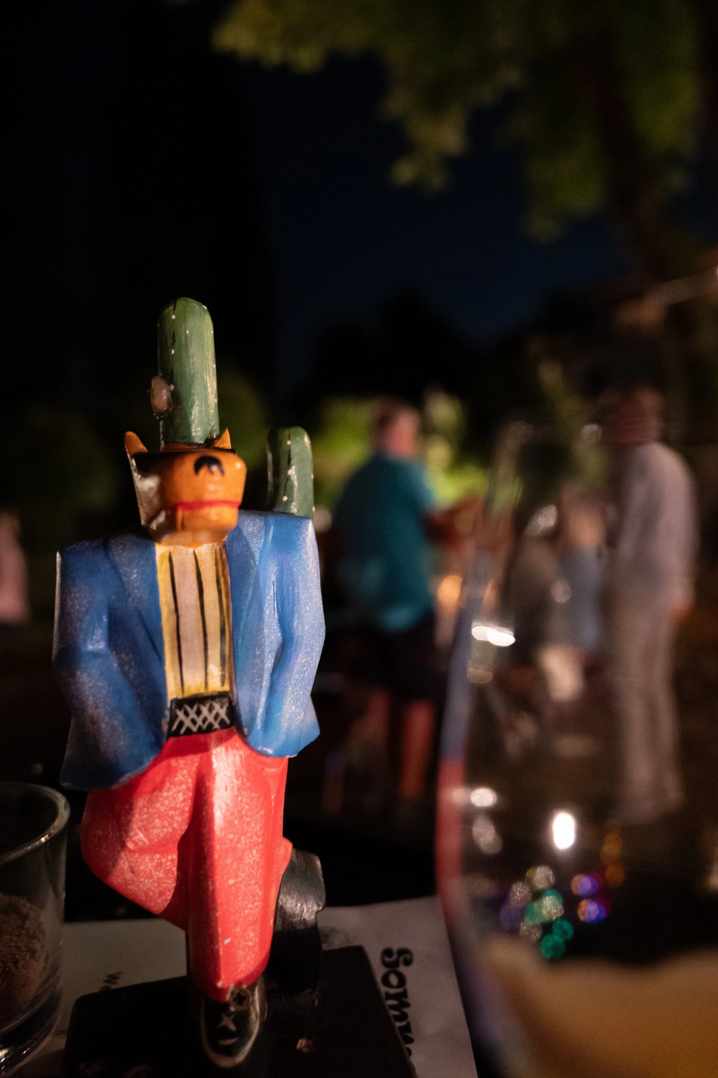 Ein bunter, handgeschnitzter Holzfigur eines Mannes im traditionellen Outfit mit roten Hosen, blauer Jacke, gelbem Hemd, grüner Mütze und schwarzem Bart. Im Hintergrund sind verschwommen Menschen bei Nacht, bei einer Outdoor-Veranstaltung oder einem 