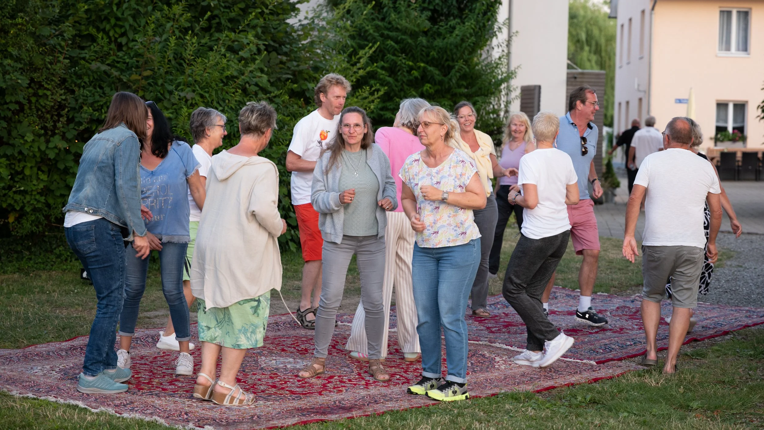 Mehrere Menschen tanzen auf einem roten Teppich im Freien, umgeben von Büschen und Gebäuden.