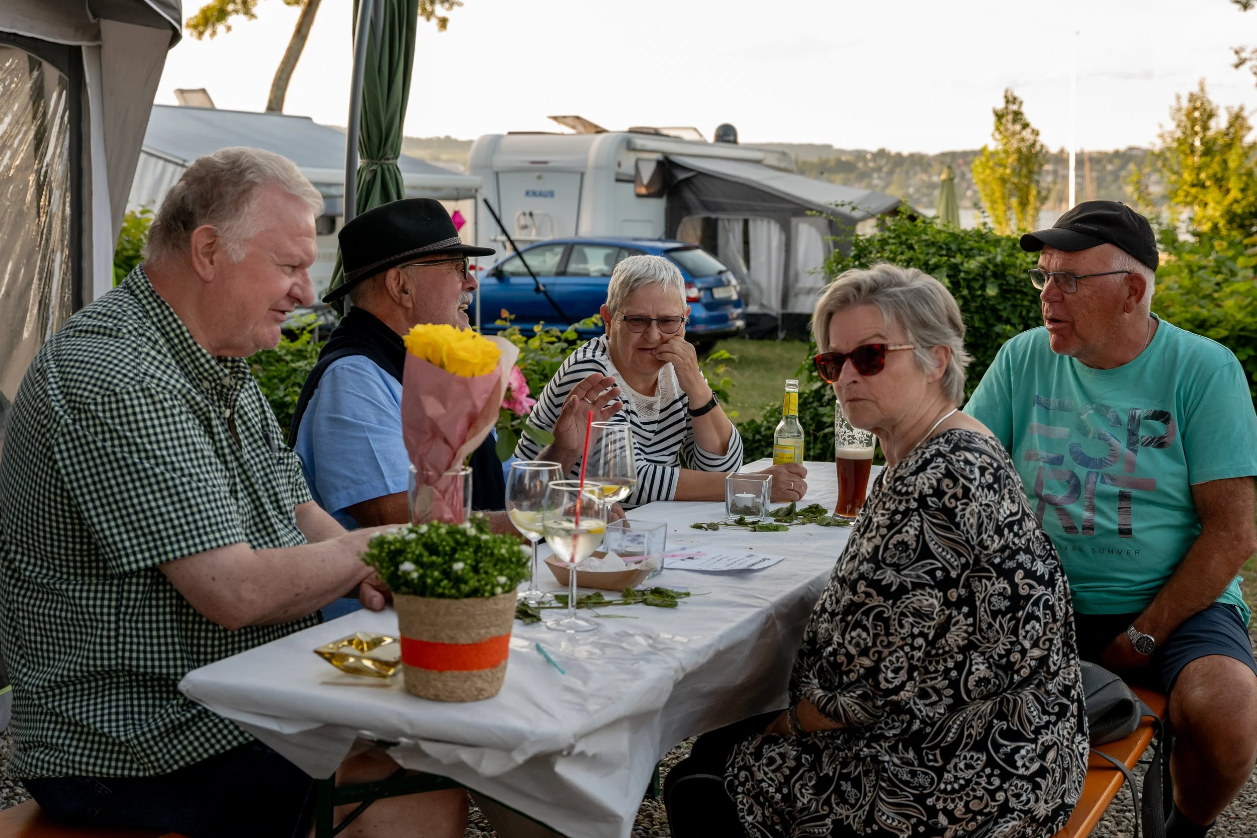 Sechs ältere Menschen sitzen bei einem Picknick im Freien an einem Tisch mit Getränken, Blumen und Papieren, während sie sich unterhalten und lachen.