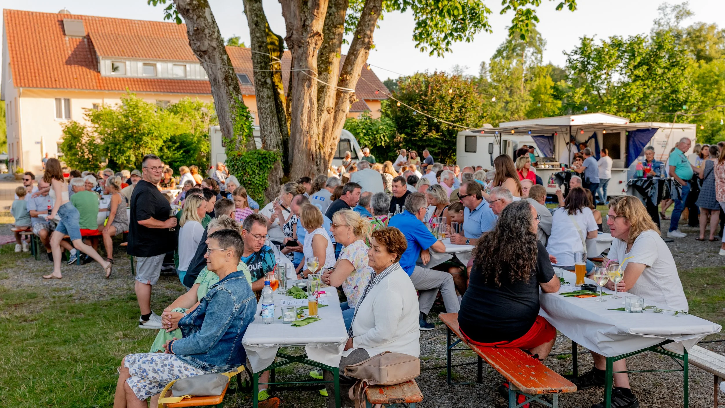 Menschen sitzen bei einem Outdoor-Gartenfest an Bierzitaten und Tafeln, im Hintergrund gibt es einen Food-Truck und Bäume