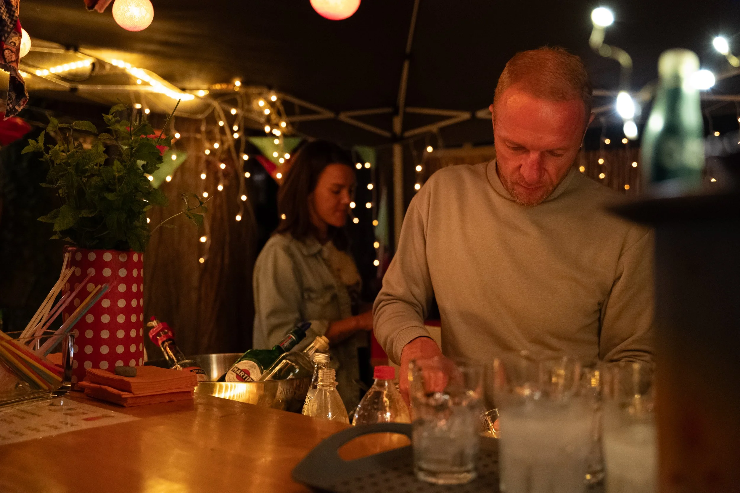 Ein Mann bereitet Getränke an einer Bar vor, im Hintergrund eine Frau und dekorative Lichter, stimmungsvolle Beleuchtung bei einer nächtlichen Feier.