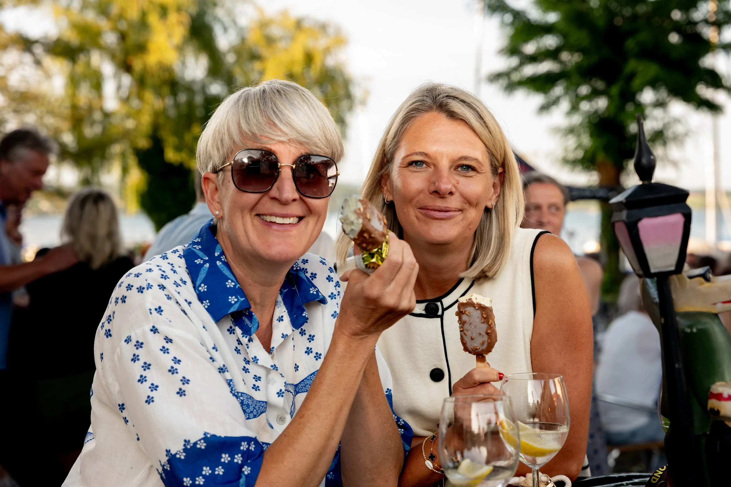 Zwei lachende Frauen genießen Eis auf einer Outdoor-Party bei Sonnenuntergang, mit Bäumen im Hintergrund.