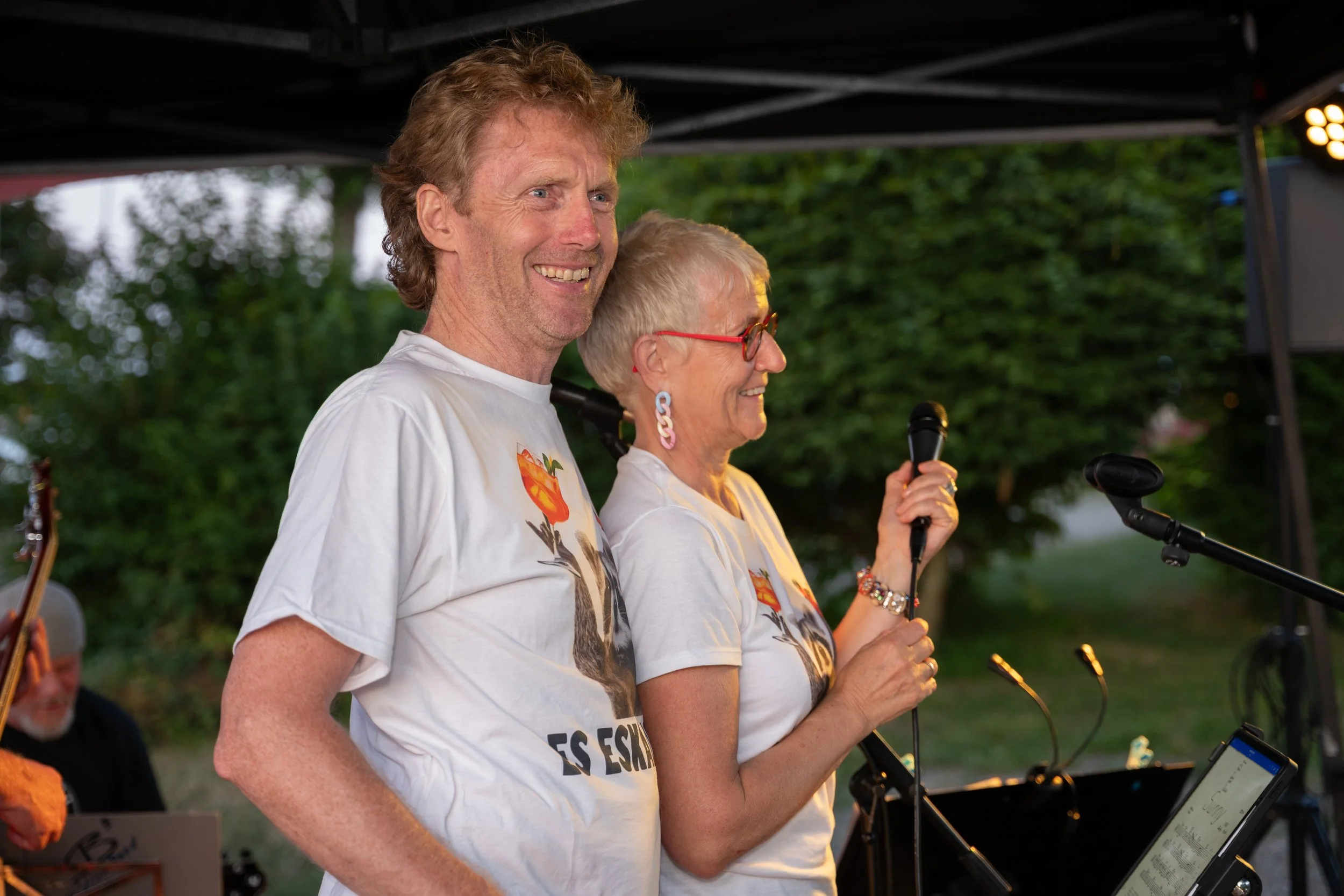 Zwei Menschen beim Singen auf einer Bühne im Freien, ein Mann und eine Frau, beide tragen weiße T-Shirts mit einem Aufdruck.