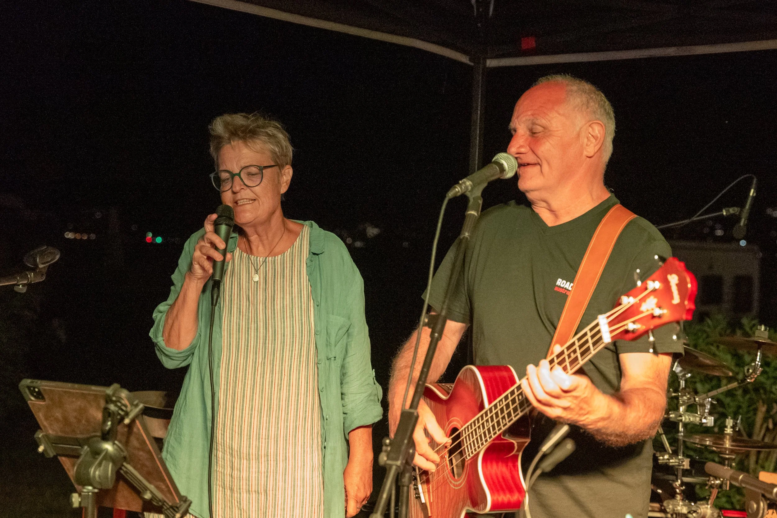 Ein älteres Paar singt und spielt Gitarre bei einer Live-Musikveranstaltung. Die Frau trägt eine grüne Jacke und die Männer ein schwarzes T-Shirt.