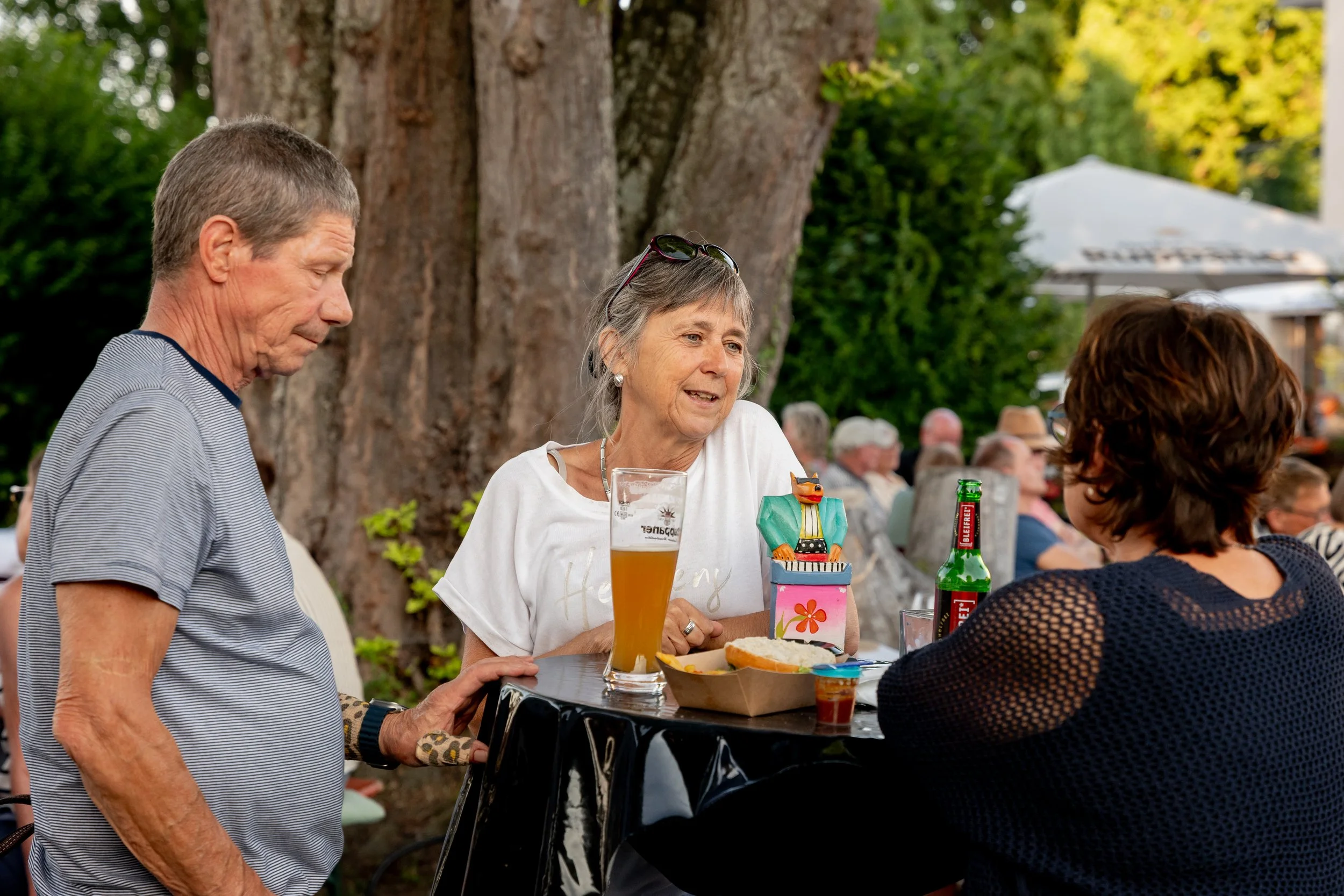 Drei ältere Menschen unterhalten sich an einem Tisch im Freien, bei einem Fest oder Event, mit großen Bäumen im Hintergrund, einem Getränk und einem Teller mit Essen auf dem Tisch.