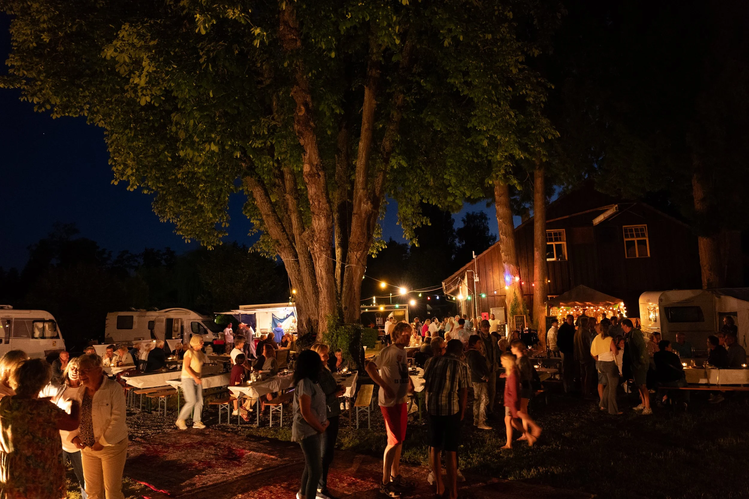 Eine nächtliche Camping-Party im Freien mit Menschen, die unter einem großen Baum sitzen und tanzen, beleuchtet von Lichterketten, neben Wohnwagen und einem Haus.