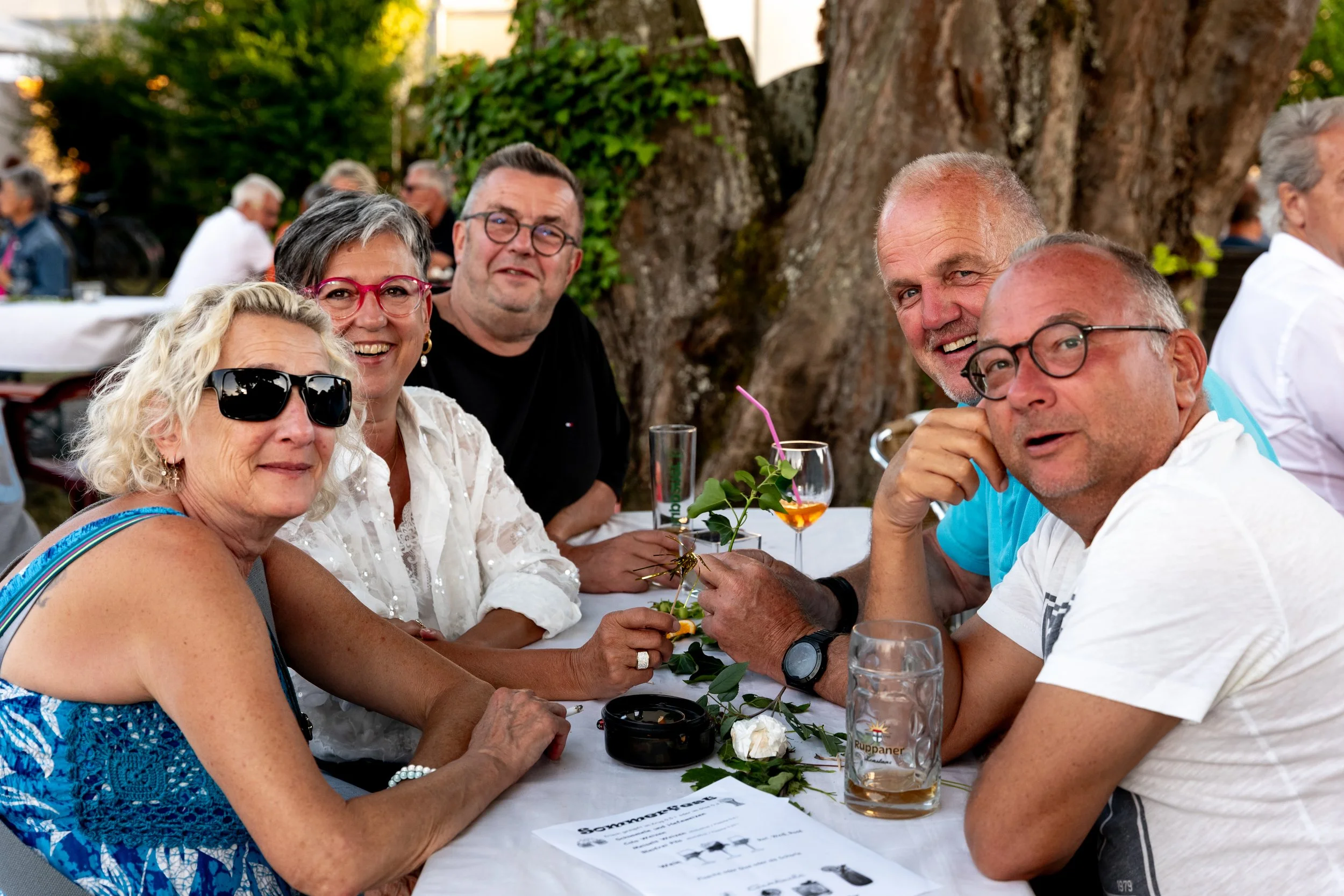 Gruppe von sechs Menschen, drei Männer und drei Frauen, sitzen an einem Tisch im Freien bei einem Treffen, lächeln und unterhalten sich. Der Tisch ist dekoriert mit Blumen und Getränken, im Hintergrund sind mehr Leute und Bäume sichtbar.