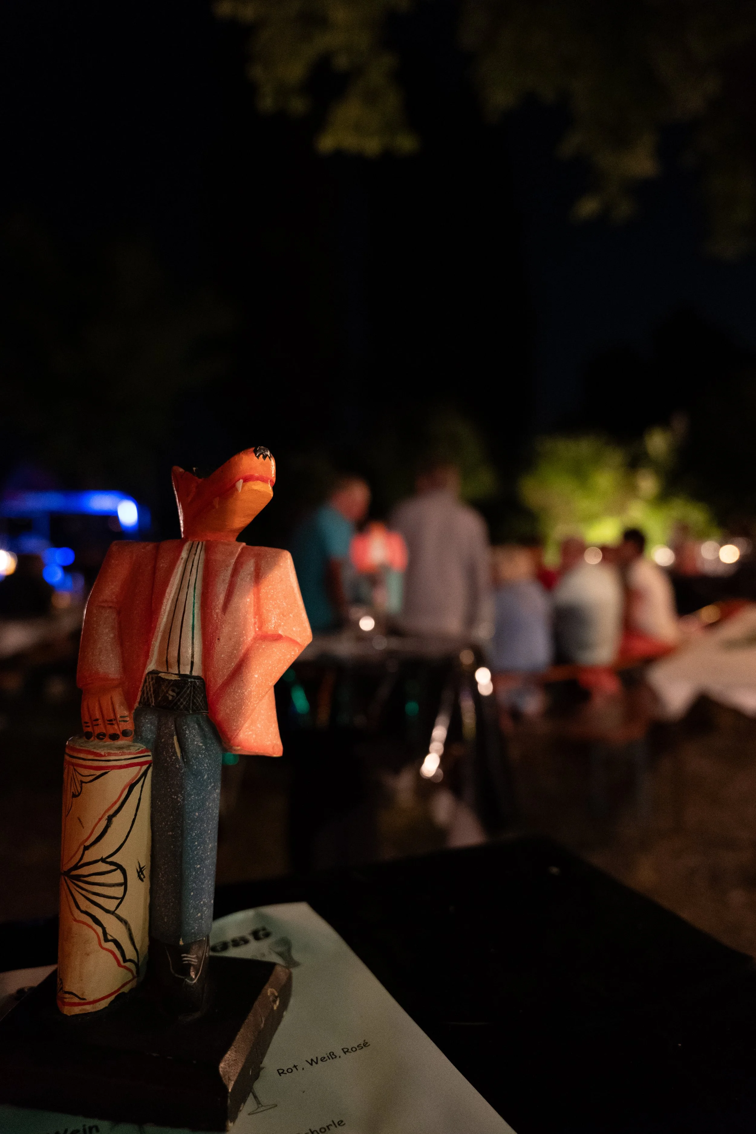 Kunststofffigur eines Sherlock Holmes mit rotem Hut und Trenchcoat, auf einem Tischtisch bei einem nächtlichen Outdoor-Event, im Hintergrund sind Gruppen von Menschen unter Bäumen sichtbar.