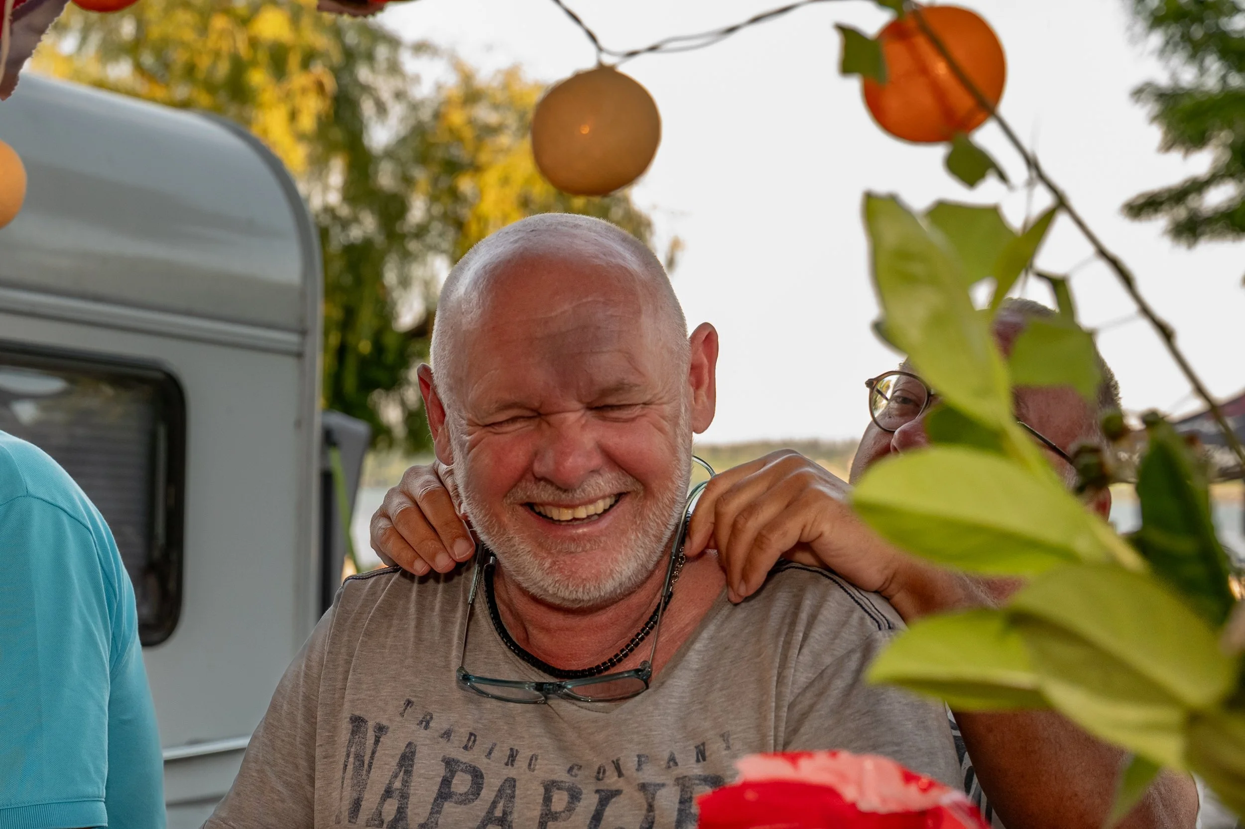 Lächelnder alter Mann mit grauem Bart, umgeben von Freunden bei einer Feier im Freien, mit Bäumen und Orangenlaternen im Hintergrund.