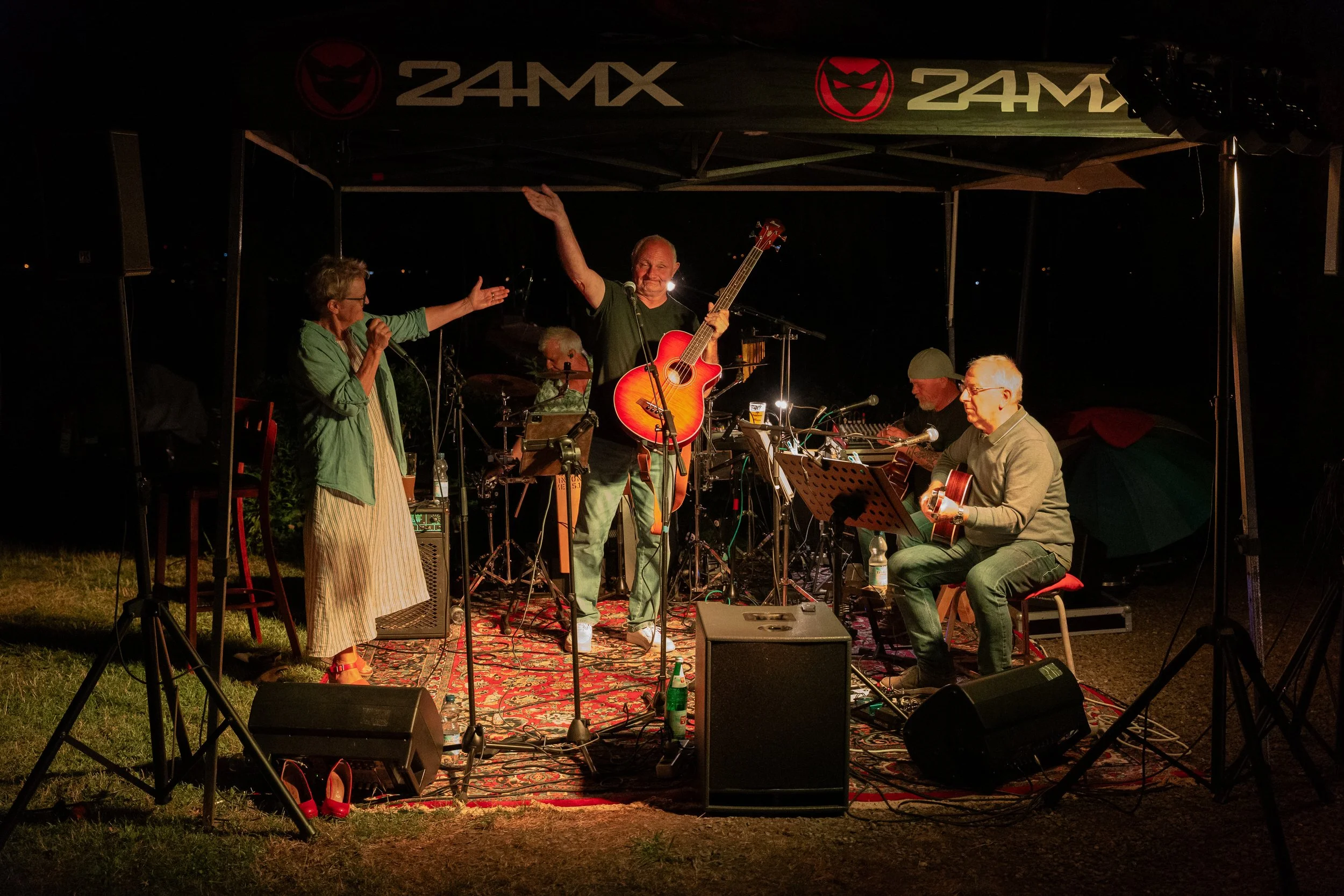 Eine Band spielt bei einem nächtlichen Outdoor-Konzert unter einem Pavillon mit der Aufschrift '24MX'. Es sind fünf Musiker zu sehen, die Gitarren und ein Schlagzeug spielen, eine Sängerin singt, während die Bühne mit Teppich ausgelegt ist.