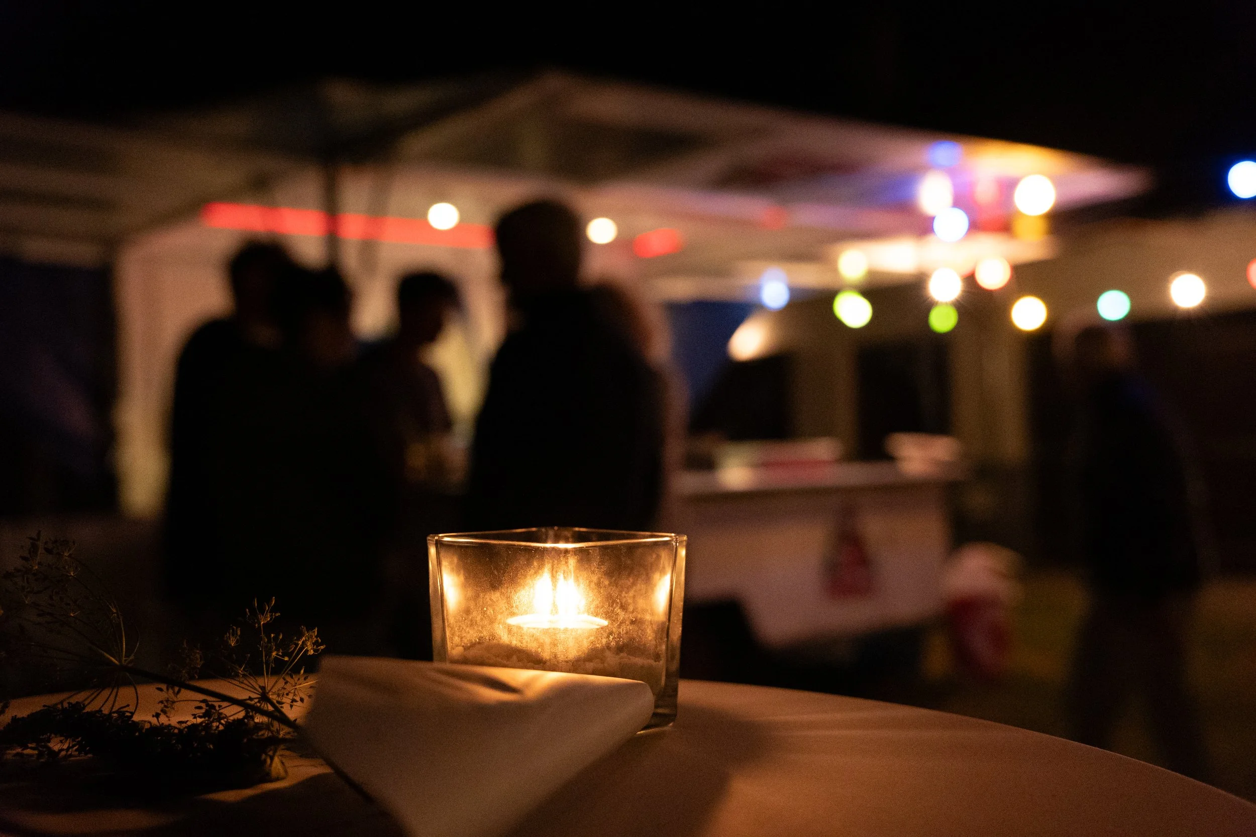Eine brennende Kerze auf einem Tisch im Vordergrund, im Hintergrund eine Gruppe von Menschen in einer dunklen, mit bunten Lichterketten beleuchteten Umgebung.