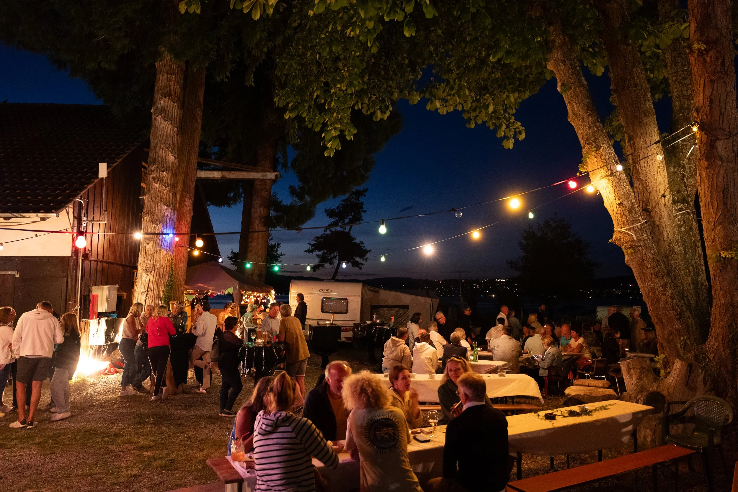 Menschen feiern abends im Freien bei einem Fest mit Lichterketten zwischen Bäumen, einige sitzen an Tischen, andere stehen und unterhalten sich, im Hintergrund sind Zelte und ein Wohnwagen zu sehen.