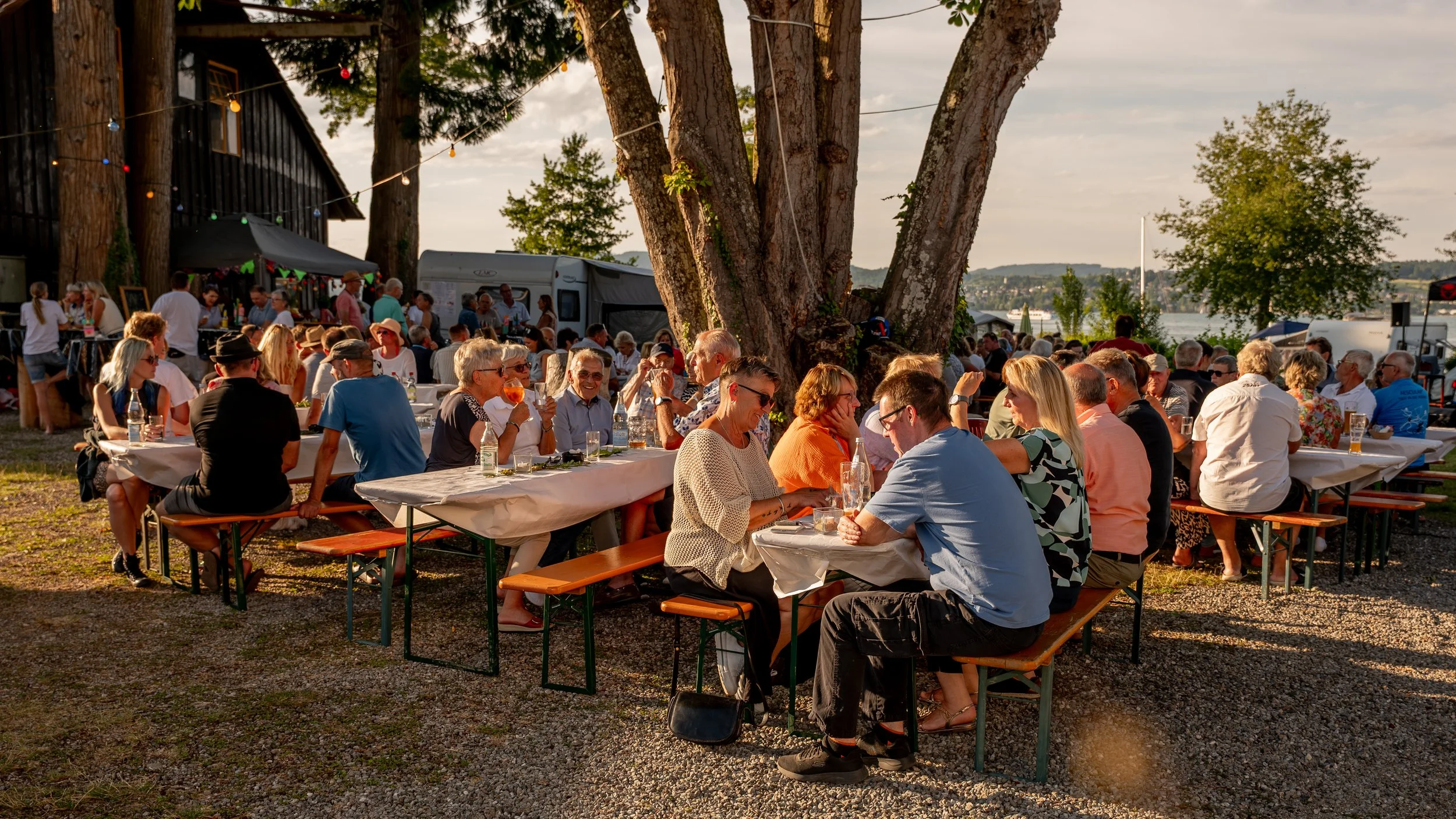 Menschen sitzen an Tischen im Freien bei einem Fest, umgeben von Bäumen, mit Wasser im Hintergrund bei Sonnenuntergang.