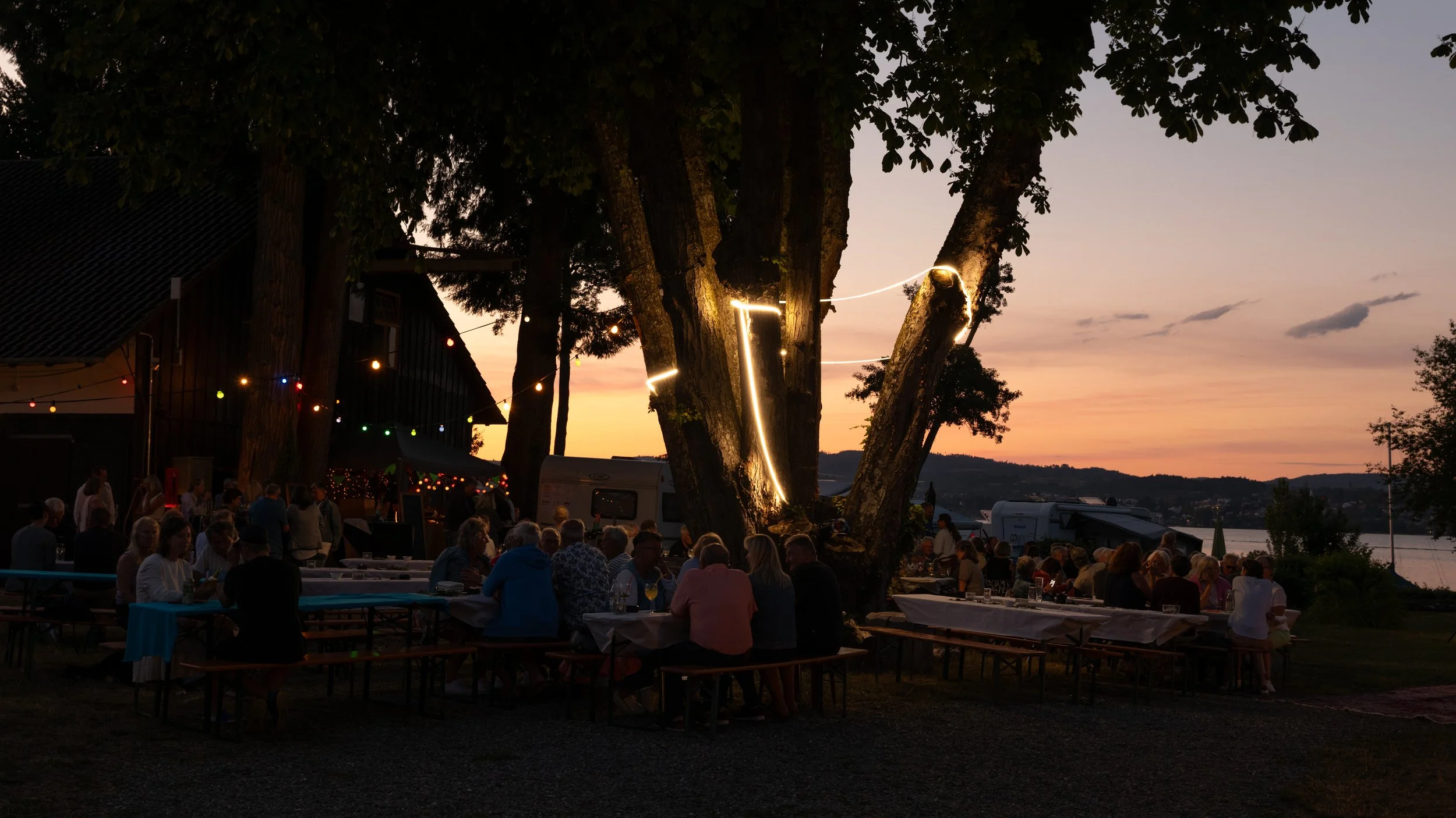 Ein Abendpicnic im Freien mit vielen Menschen, die an Tischen sitzen, während die Sonne untergeht. Im Hintergrund sieht man Wasser und Berge, dekorierte Lichter hängen zwischen den Bäumen.