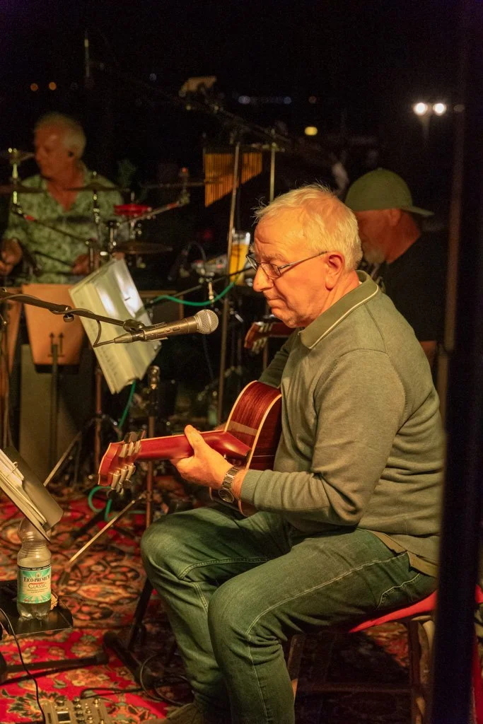 Ein älterer Mann spielt Gitarre während eines Live-Musikauftritts in einem dunklen Raum.