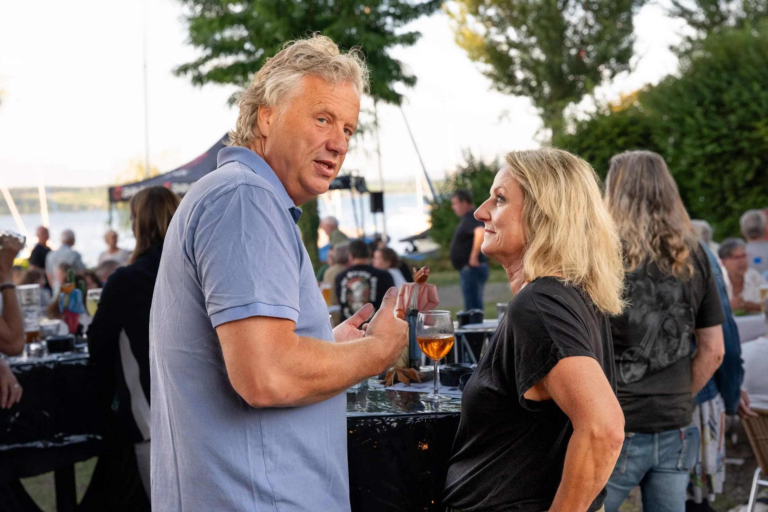 Ein Mann und eine Frau unterhalten sich bei einem Outdoor-Event, im Hintergrund sind weitere Personen, Wasser und Bäume sichtbar.