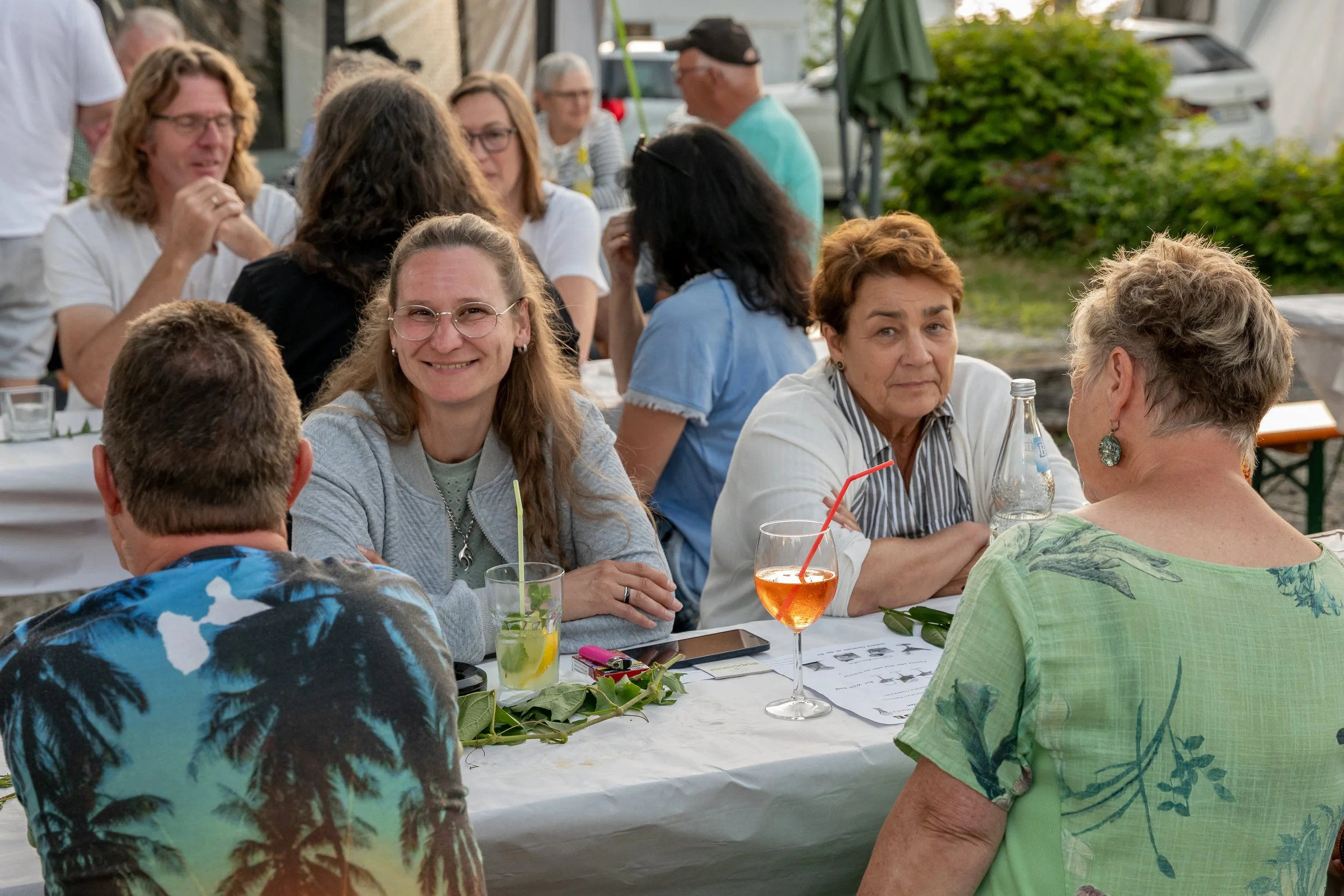 Menschen sitzen an einem länglichen Tisch bei einer Feier im Freien, trinken Getränke, unterhalten sich, im Hintergrund sind weitere Gäste und grüne Pflanzen sichtbar.