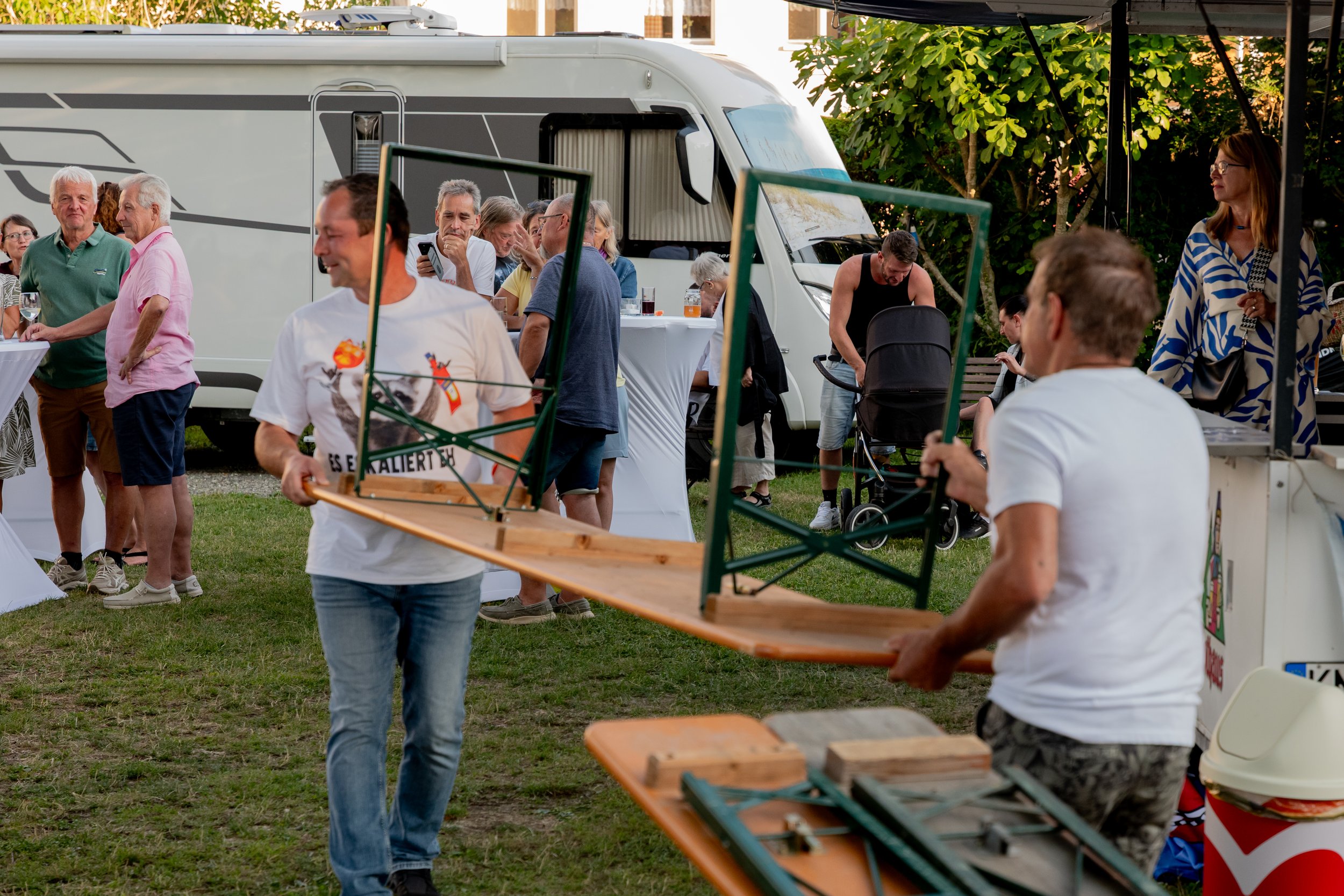 Menschen auf einem festivalähnlichen Event, einer Person bringt ein großes Holzgestell. Im Hintergrund stehen Leute beim Essen und eine Person schiebt einen Kinderwagen. Es gibt Zelte und einen Wohnwagen.