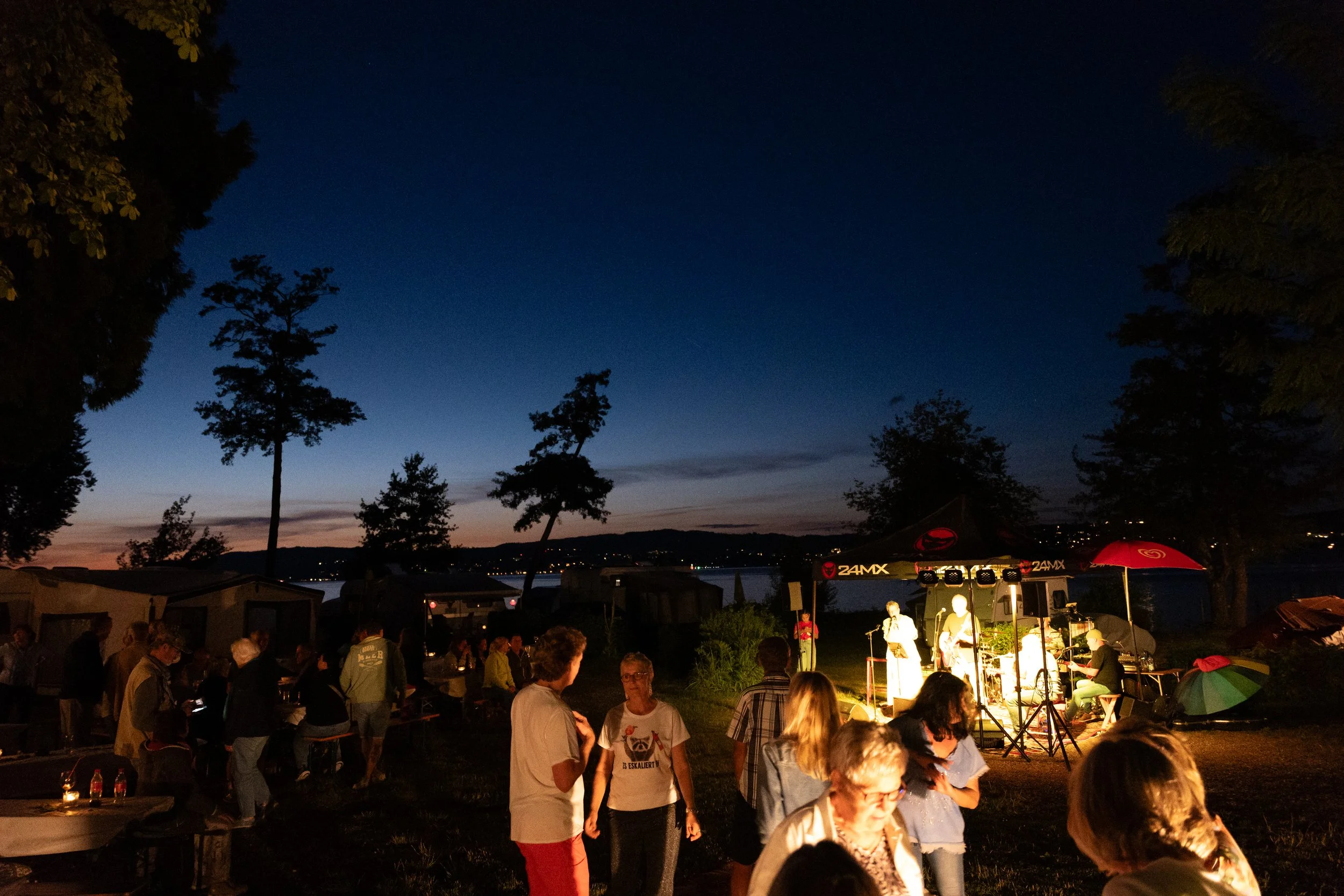 Ein nächtliches Bild eines Outdoor-Musikkonzerts mit einer Bühne, auf der Musiker auftreten, umgeben von Menschen, einige im Gespräch, andere beim Zuschauen. Es gibt Sonnenschirme, Bäume und einen See im Hintergrund, der Himmel ist dunkelblau.