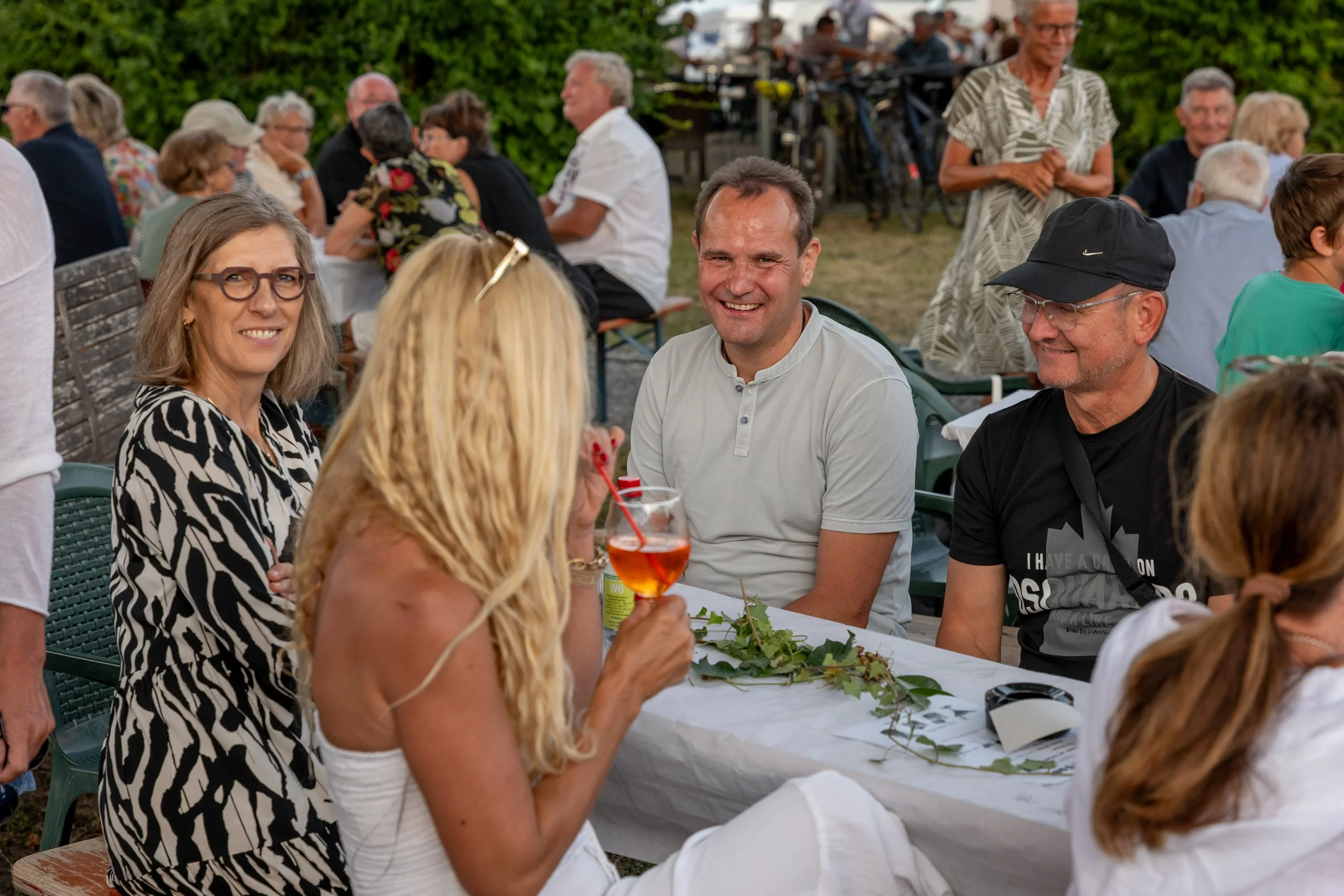 Menschen sitzen bei einer Party im Freien, trinken Getränke und unterhalten sich, im Hintergrund sind weitere Gäste und Fahrräder zu sehen.