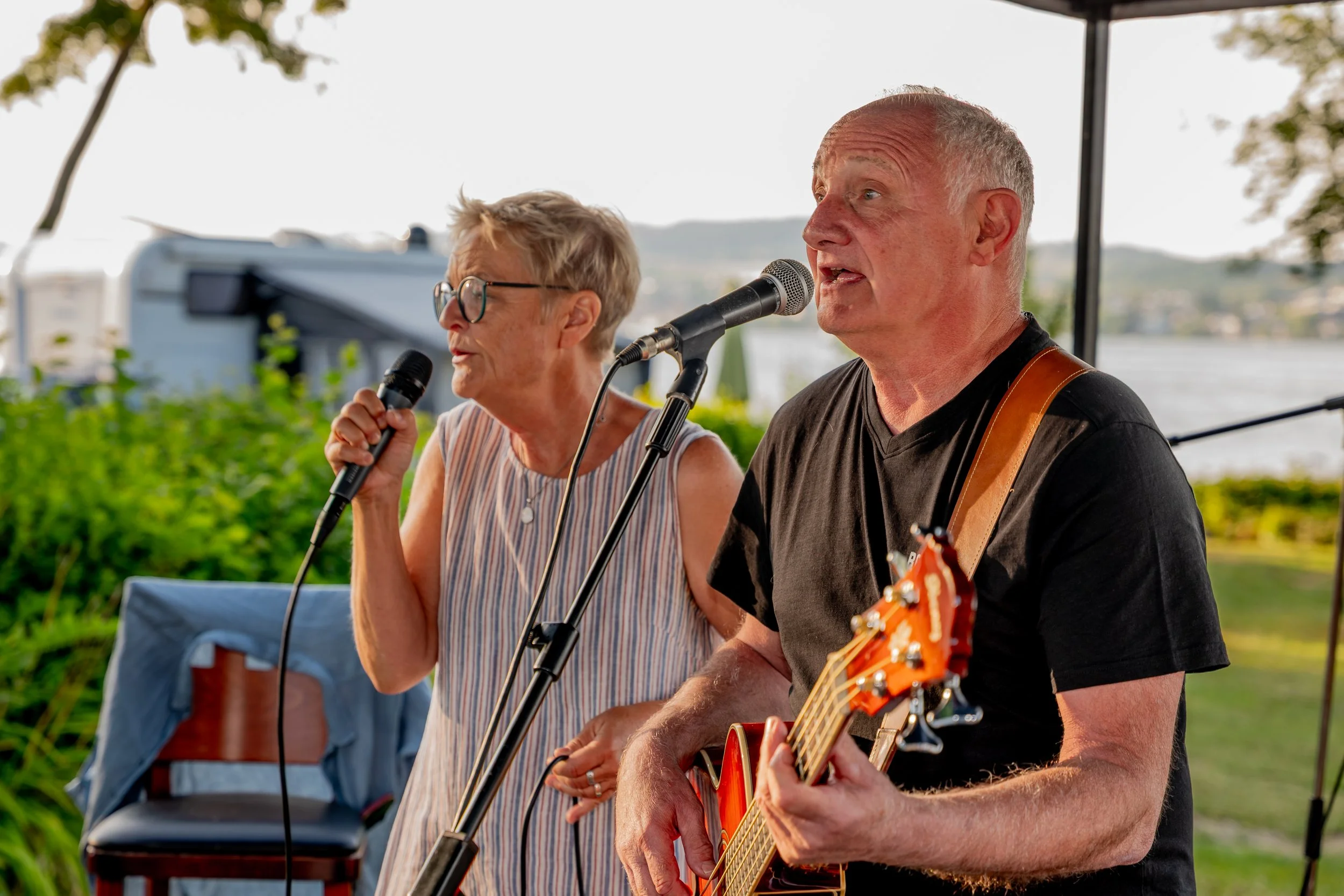 Ein älteres Paar singt und spielt Gitarre bei einem Open-Air-Konzert am Wasser, im Hintergrund sind Bäume und ein Boot sichtbar.
