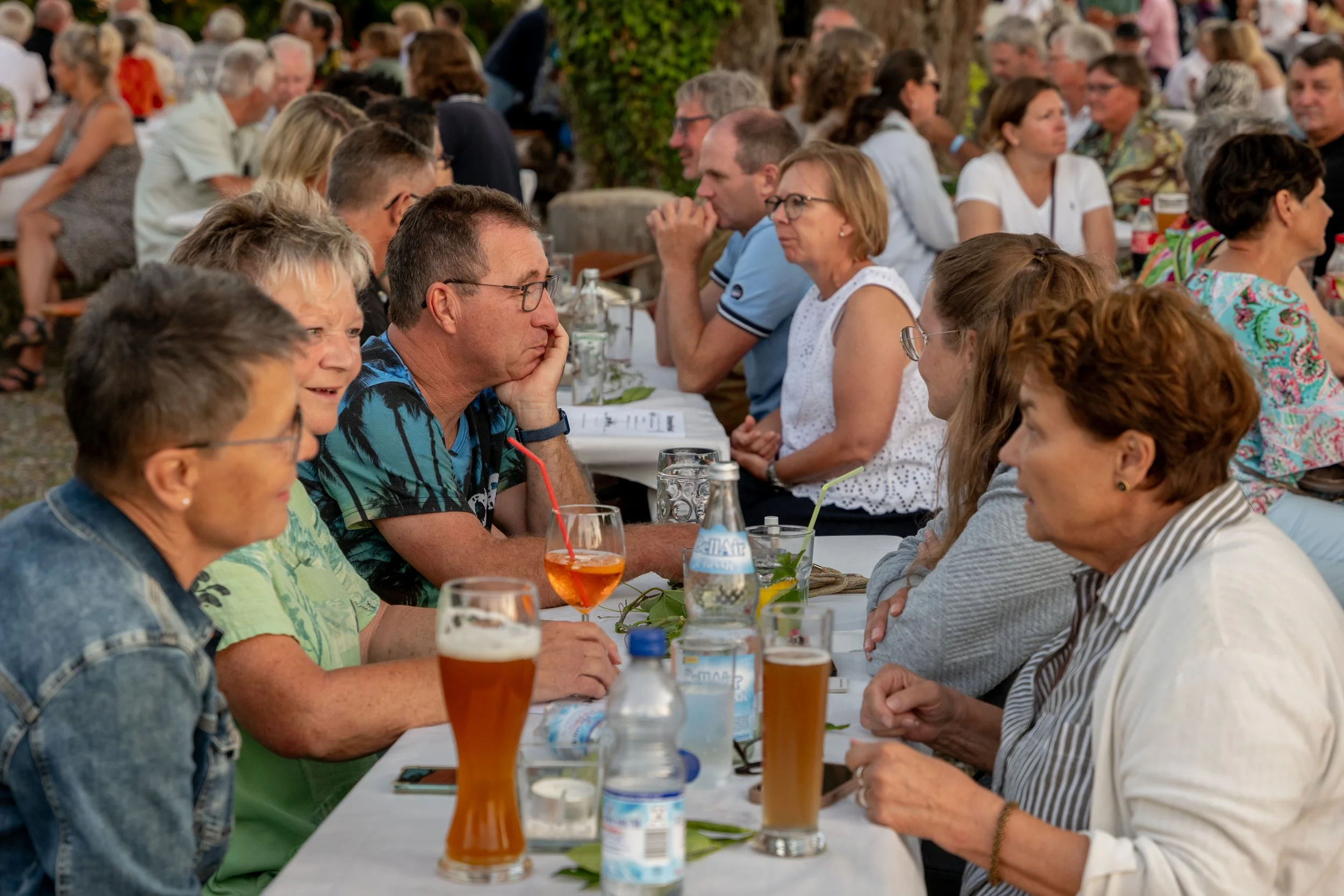Menschen sitzen an einem langen Tisch bei einer Feier im Freien, trinken Bier und unterhalten sich.