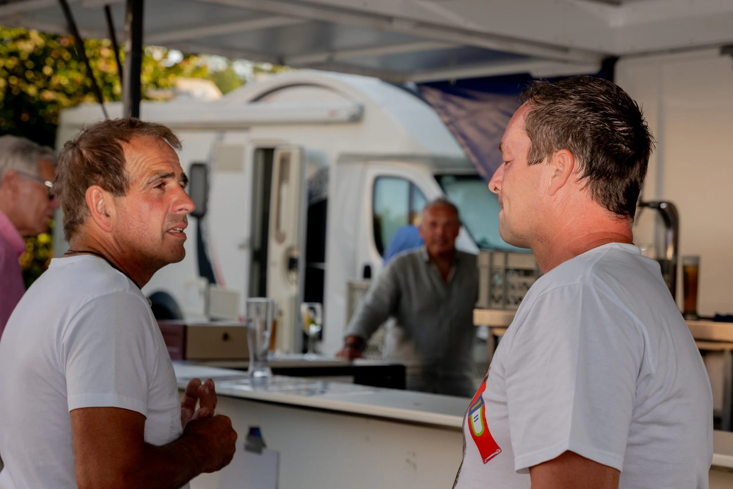 Zwei Männer im Gespräch vor einem Food-Truck bei einem Festival oder Markt, im Hintergrund ein weiteres älteres Mann. Es ist sonnig, und die Männer tragen weiße T-Shirts.
