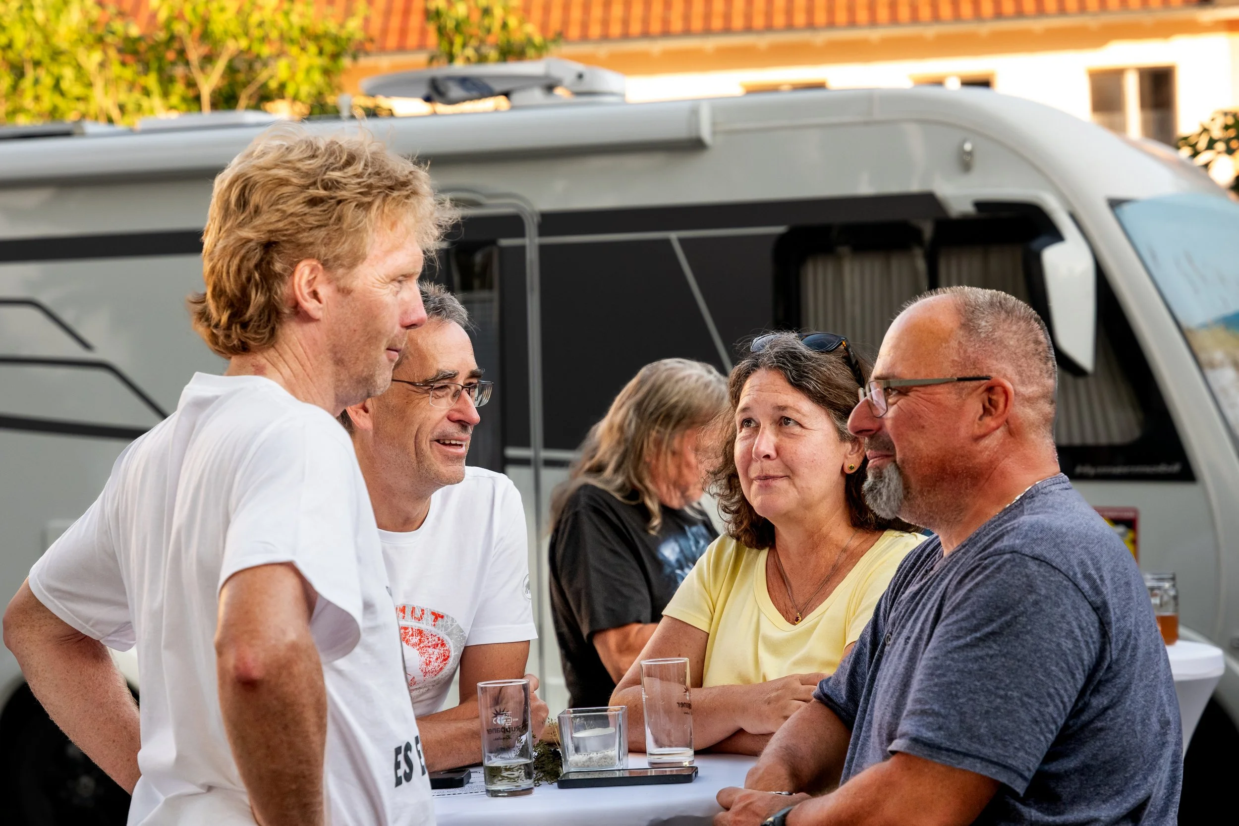 Menschen, die bei einem Treffen im Freien um einen Tisch sitzen, unterhalten sich, mit einem Camper im Hintergrund.
