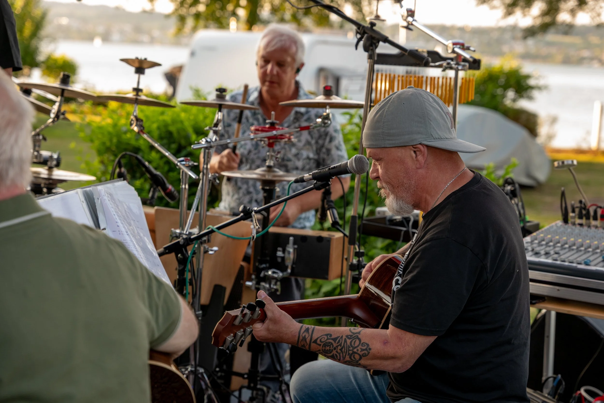 Musiker spielen im Freien bei Sonnenuntergang, einer spielt Gitarre, die anderen mit elektronischen Instrumenten, im Hintergrund Wasser und Bäume.
