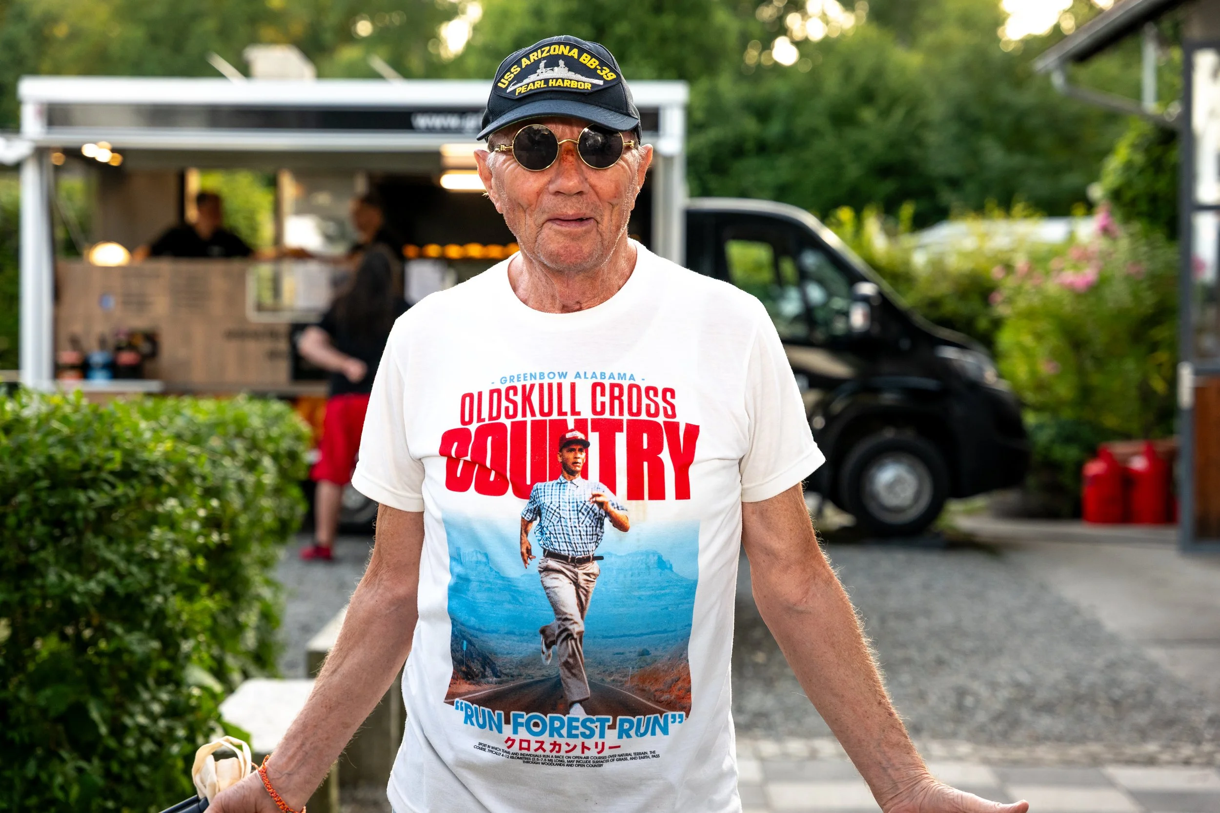 Älterer Mann mit Sonnenbrille und Baseballmütze vor Food-Truck, trägt T-Shirt mit Laufmotivi, im Hintergrund grüne Bäume und eine schwarze Lieferwagen.