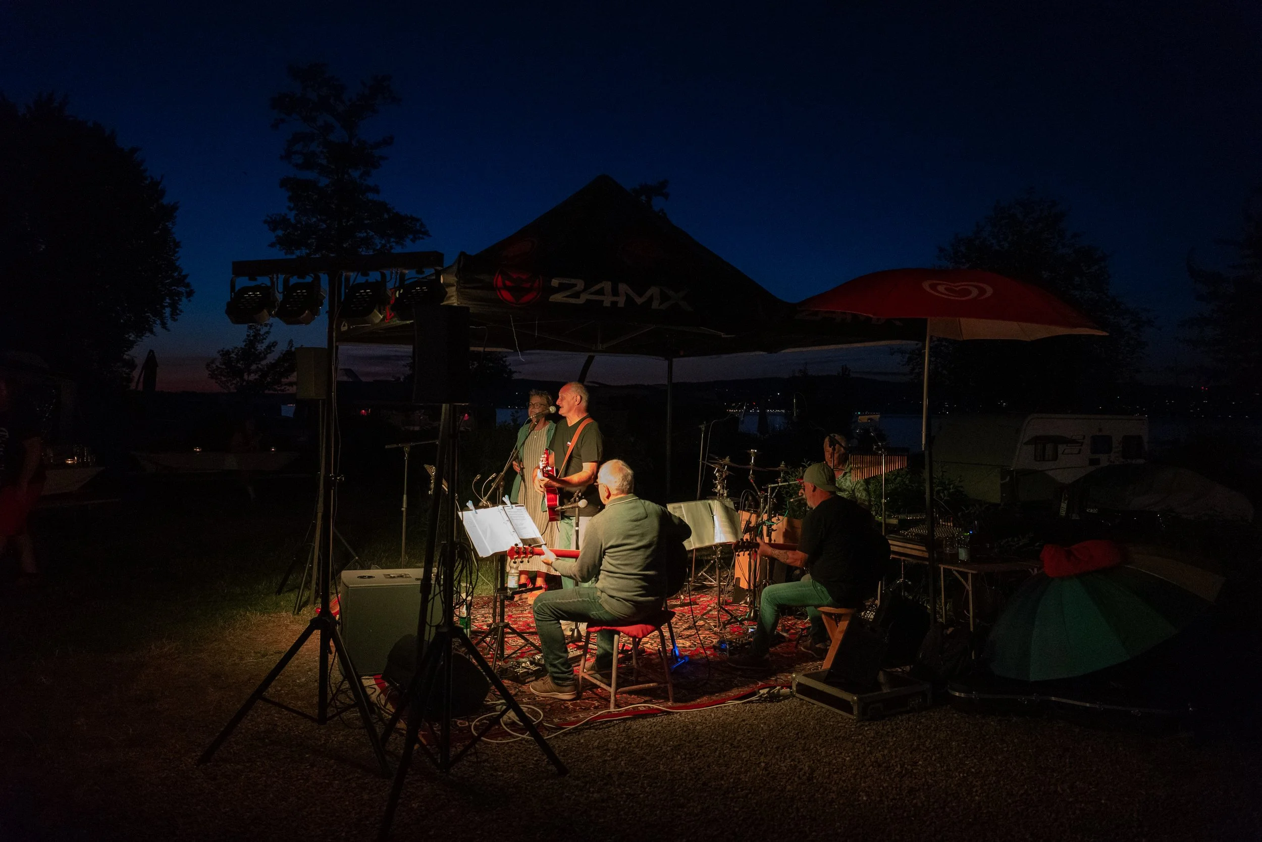 Musikgruppe spielt im Freien bei Nacht unter Zelten, mit Bäumen im Hintergrund.