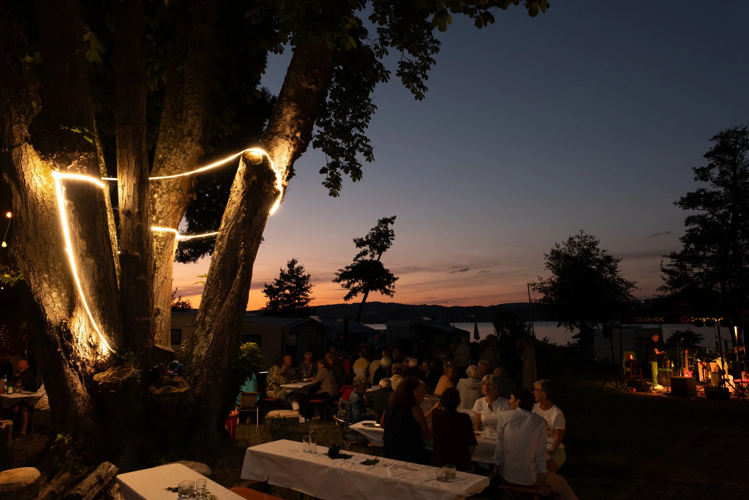 Geselliges Treffen im Freien bei Sonnenuntergang, viele Menschen sitzen an Tischen, ein Baum mit Lichterketten im Vordergrund, Wasser und Hügel im Hintergrund.