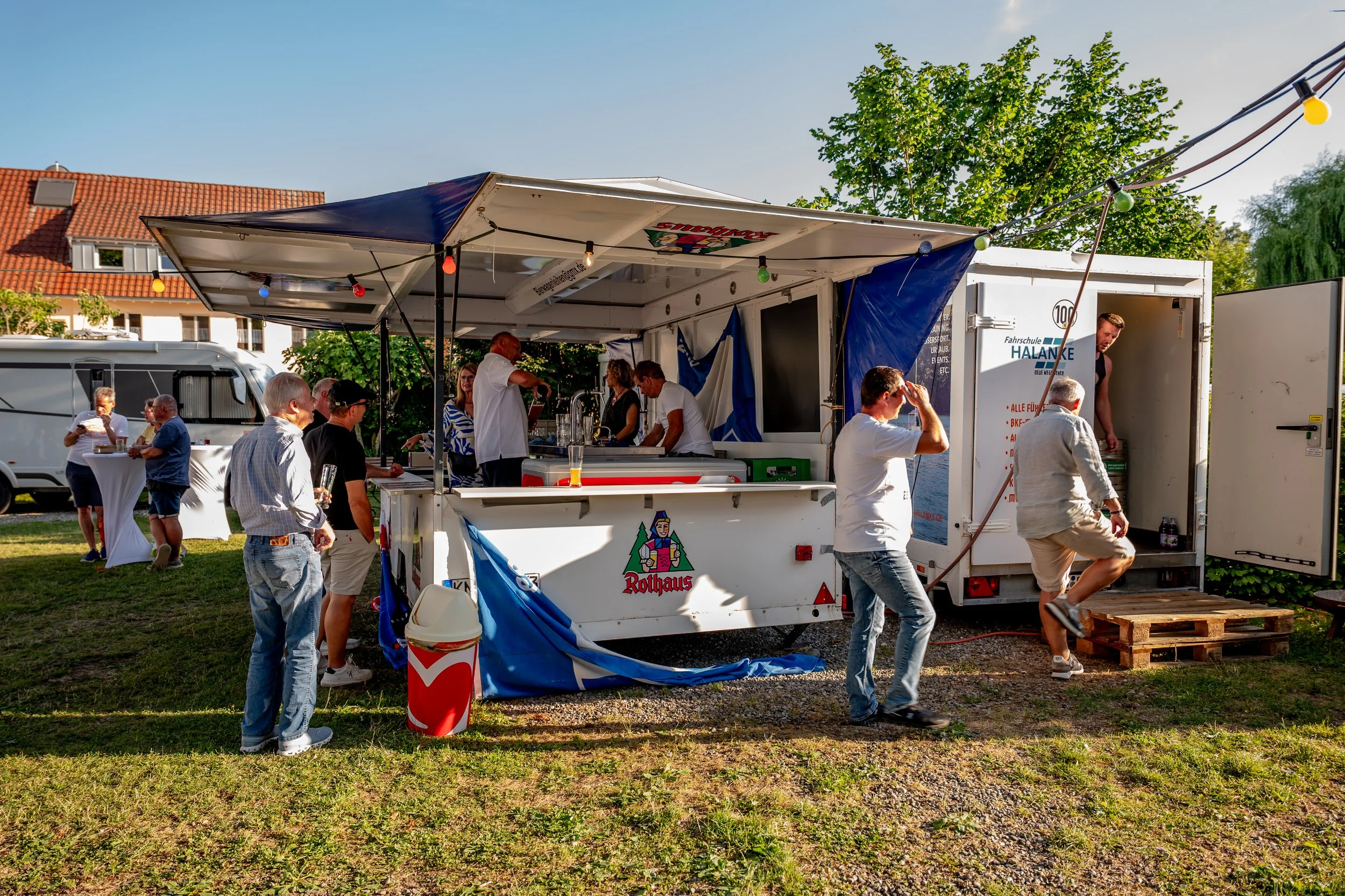 Menschen feiern um einen mobilen Bierverkaufstand im Freien, mit bunten Lichtern, im Sonnenuntergang, auf einem Gras- und Kiesboden, bei einem Fest in Deutschland.