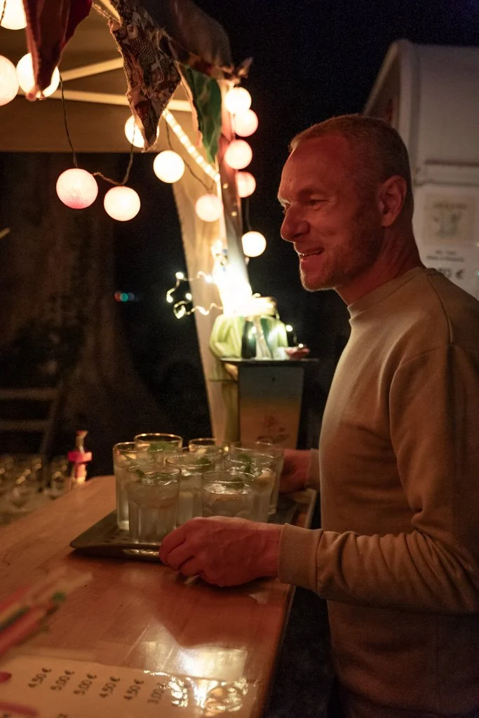 Ein Mann steht an einem improvisierten Barstand bei Nacht, umgeben von Lichterketten, und trägt eine Platte mit mehreren Gläsern Wasser.