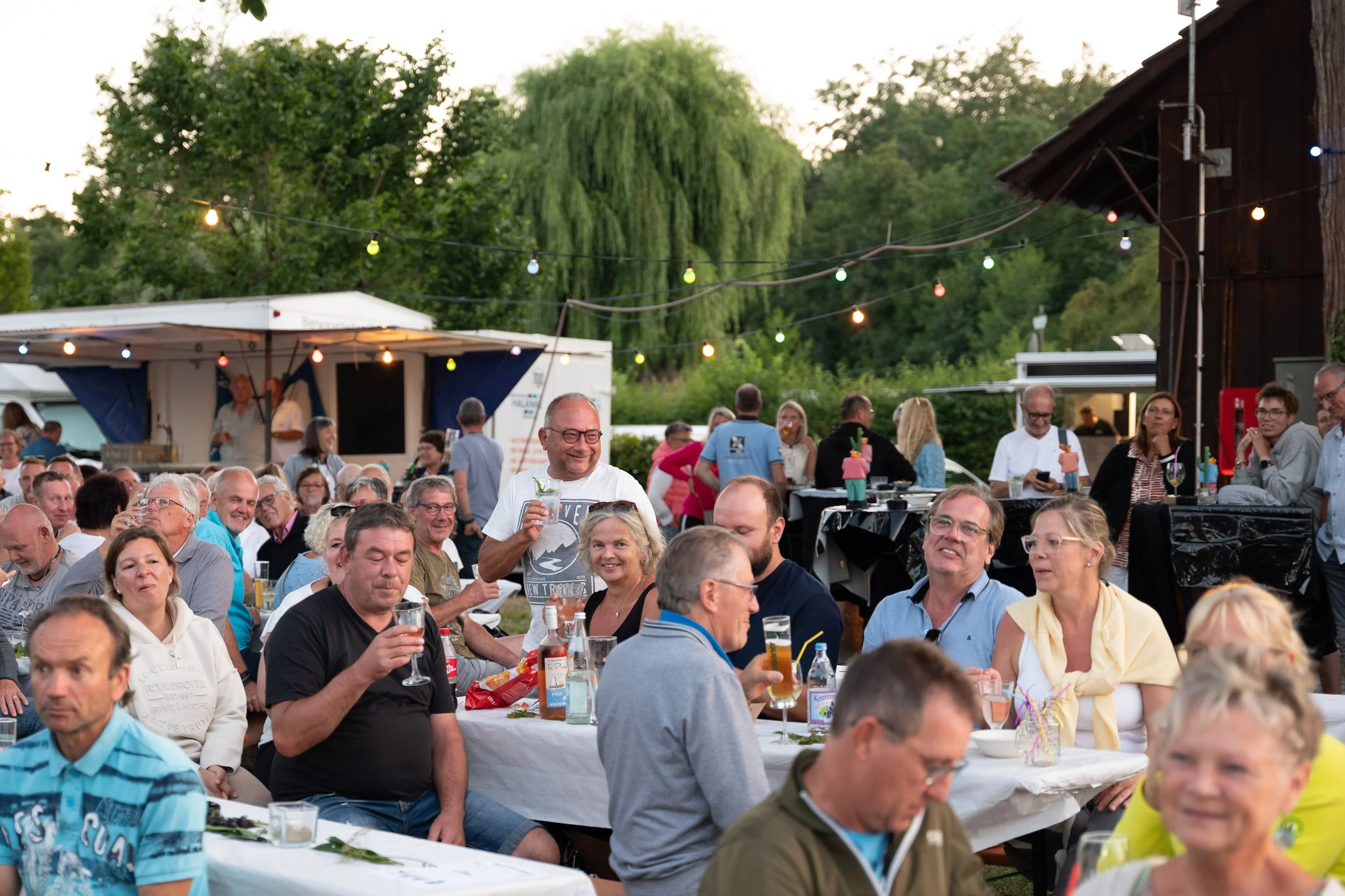 Menschen beim Fest im Freien, sitzen an Tischen, trinken und lachen, umgeben von Lichterketten, im Hintergrund Bäume und eine Food-Truck.