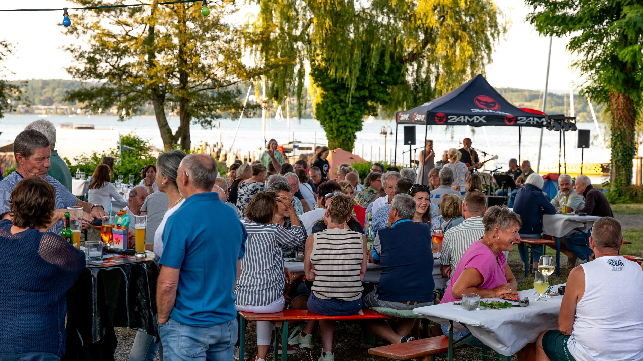 Eine Gruppe von Menschen genießt ein Open-Air-Konzert am Wasser bei Sonnenuntergang, mit Tischen, Bänken, einer Band auf der Bühne und festlicher Beleuchtung.
