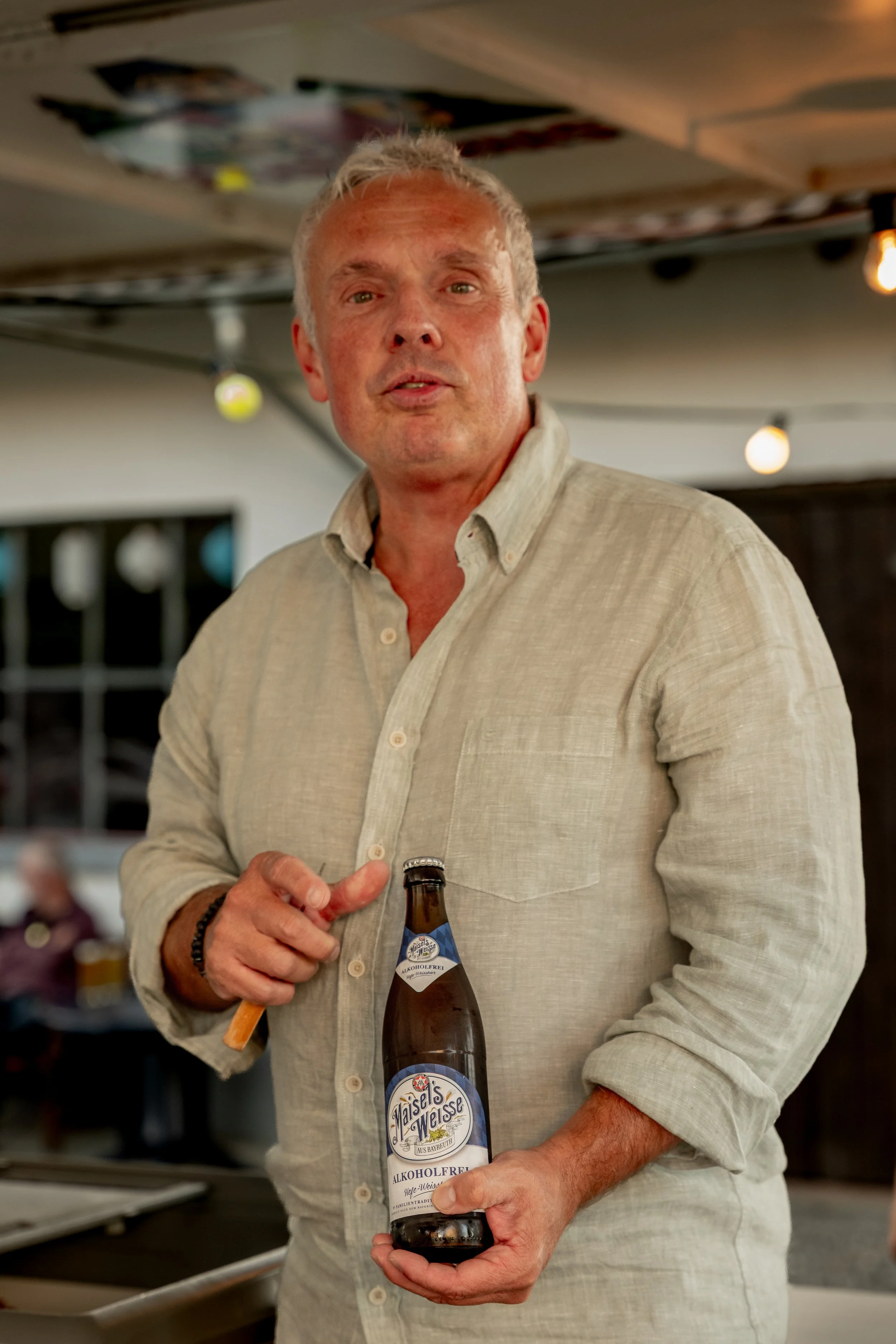 Ein Mann in einem hellen Hemd hält eine alkoholfreie Bierflasche und zeigt auf sie, während er in einem Restaurant oder einer Bar steht.