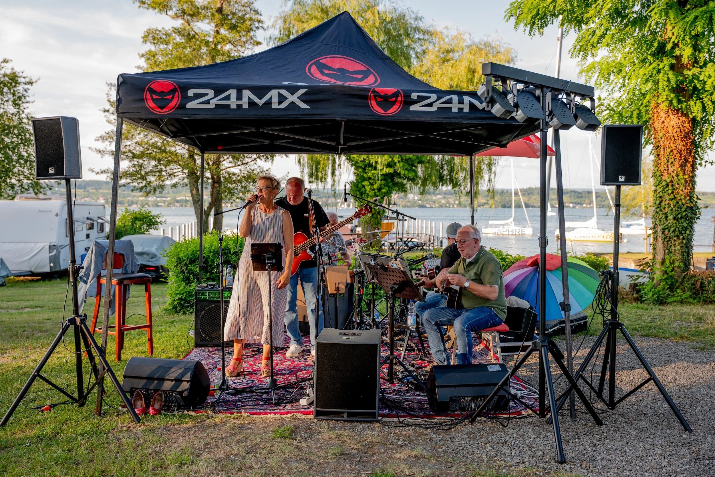 Musiker spielen auf einer Open-Air-Bühne am Wasser mit Bäumen im Hintergrund.