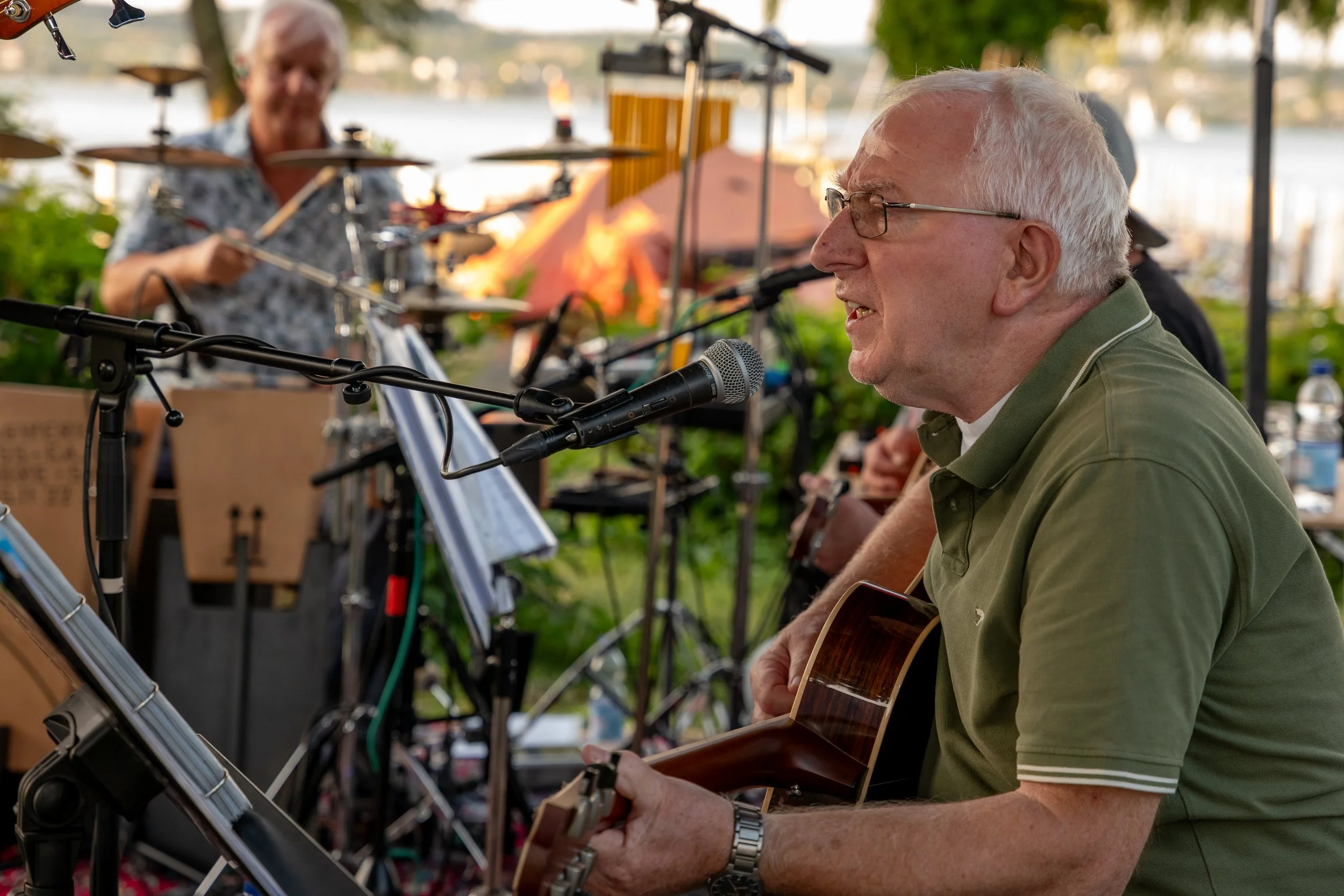 Ein älterer Mann spielt Gitarre und singt in einem Outdoor-Setting, während im Hintergrund eine Person an einem Schlagzeug spielt.